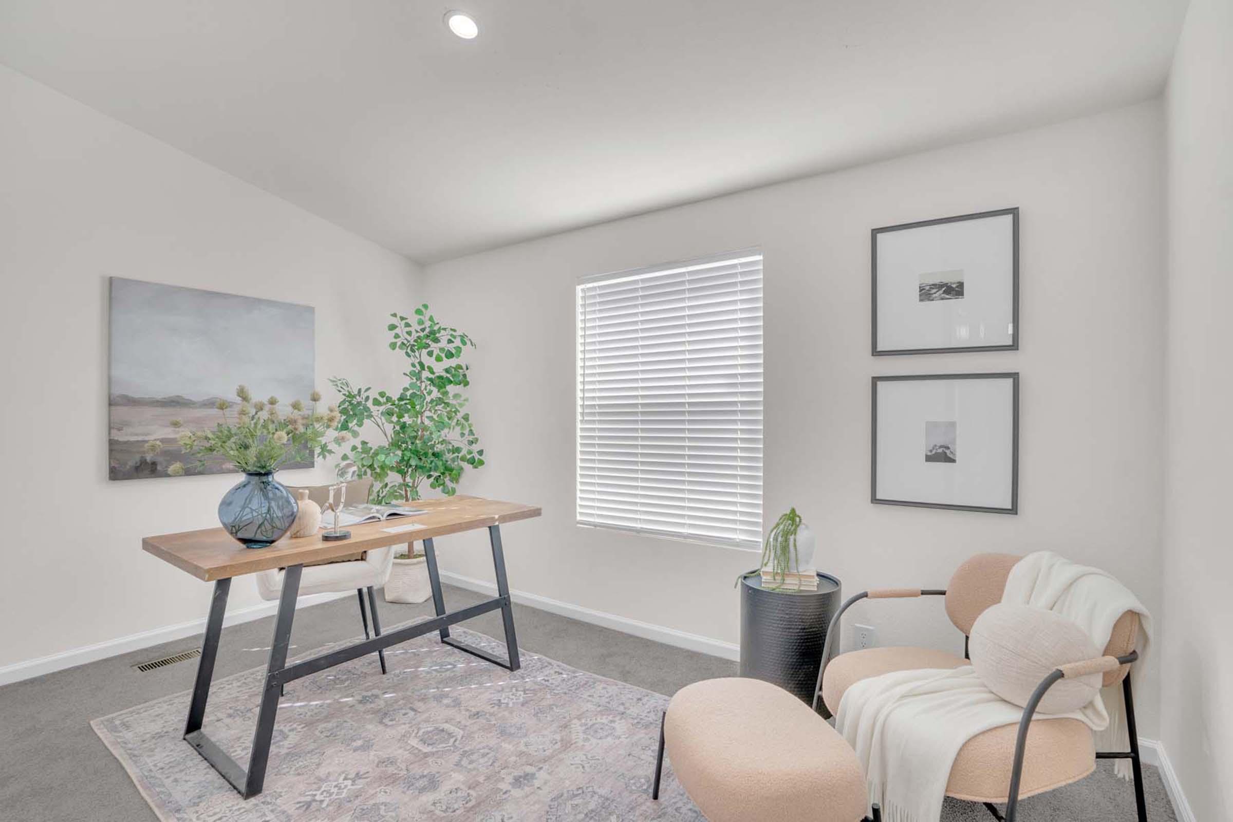 A bright, modern room featuring a wooden table with a floral arrangement, a cozy chair, and a round side table. The walls are adorned with framed artwork, and natural light streams in through a window with blinds. A soft rug adds warmth to the space, creating a welcoming atmosphere.