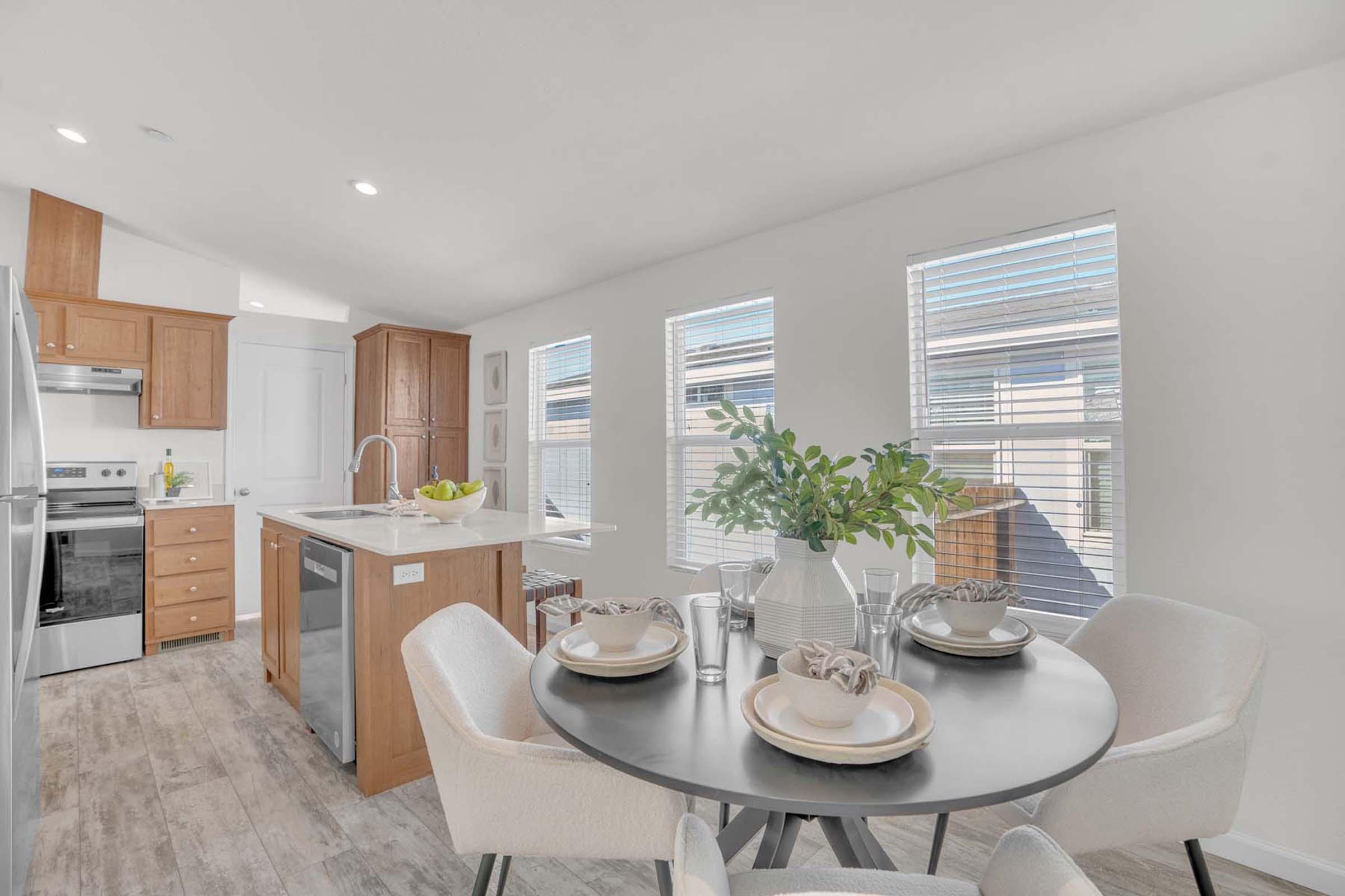 Modern kitchen dining area featuring a round black table set with plates and glasses. Light wood cabinets and stainless steel appliances complement the space. Sunlight streams through several windows with blinds, illuminating the room, which has a clean and contemporary aesthetic.