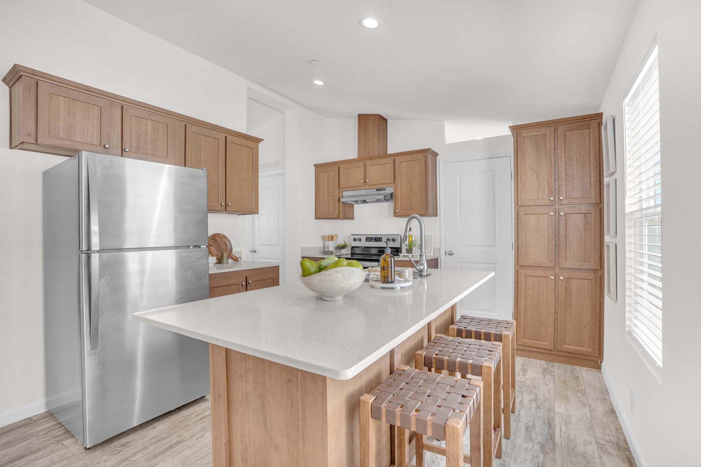 A modern kitchen featuring wooden cabinetry, a stainless steel refrigerator, a central island with a white countertop, and three wooden stools. The space is bright with natural light from a window, and there are decorative items such as a bowl of fruit on the countertop.