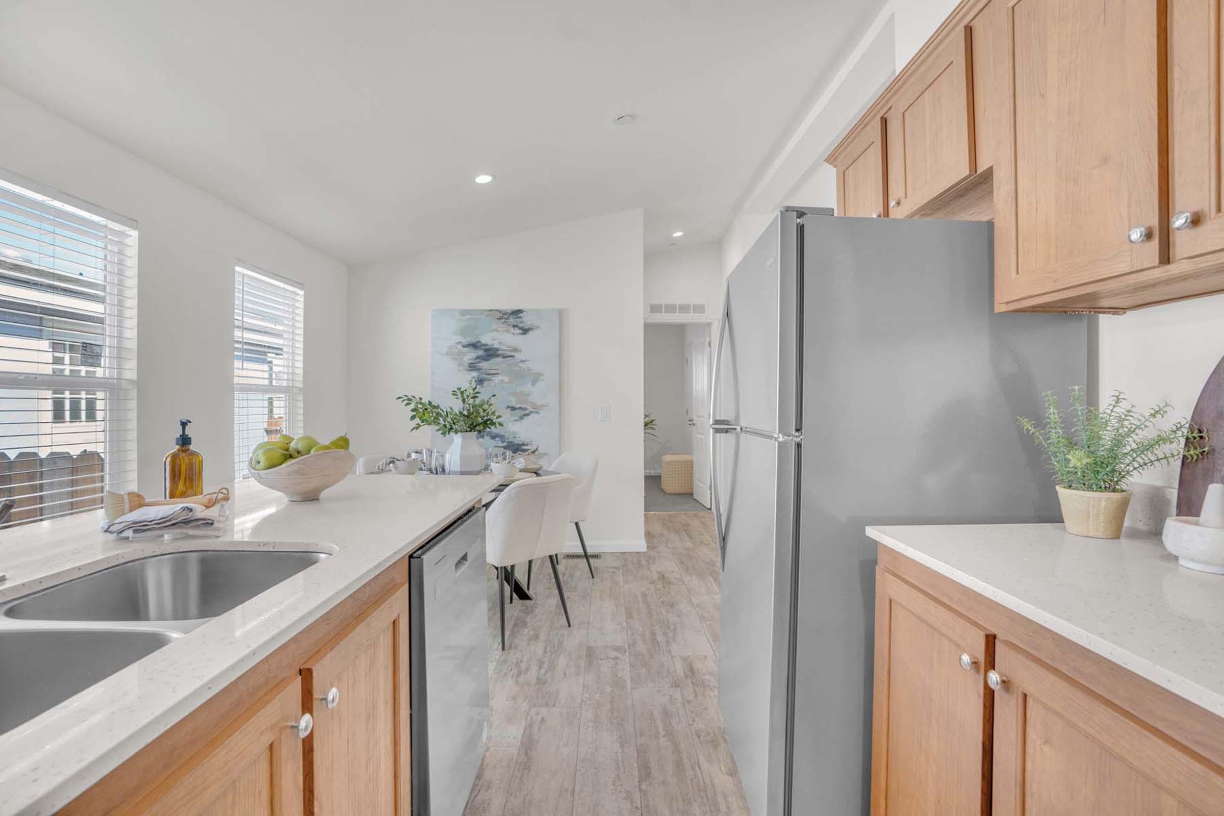 Modern kitchen featuring light wood cabinetry, stainless steel appliances, and a large sink. The space is bright with natural light from windows, and there's a dining area visible in the background. A decorative bowl with green apples and a plant add a touch of color.
