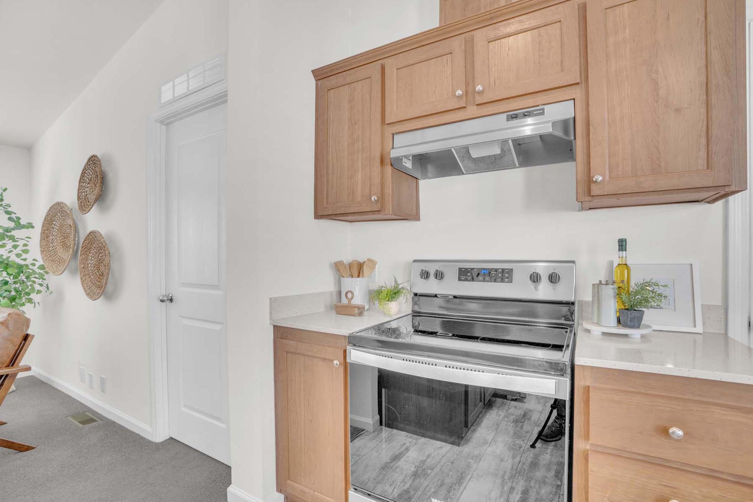 Modern kitchen corner featuring a stainless steel oven, light wood cabinetry, and a granite countertop. A few utensils are placed on the countertop, and a small plant and decorative items add a touch of greenery. The background includes a doorway leading to another room and woven wall decor.