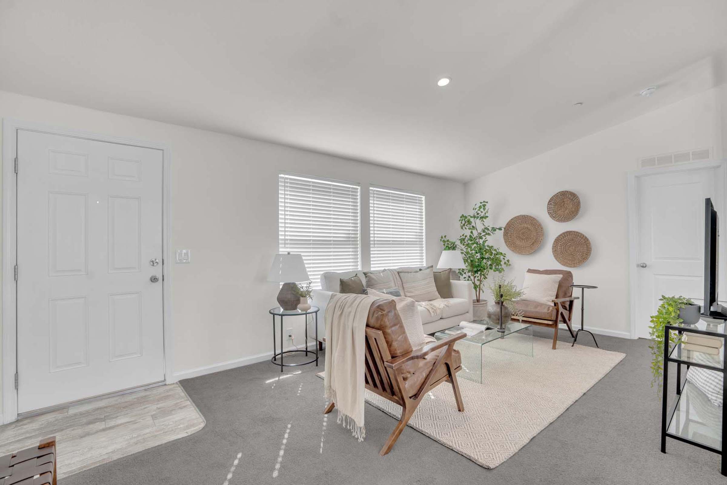 A modern living room featuring a light-colored sofa, two mid-century style armchairs, and a glass coffee table. Natural light streams through two windows with blinds. Decorative elements include a plant, a textured area rug, woven wall art, and a lamp on a side table. The walls are painted in soft neutral tones.