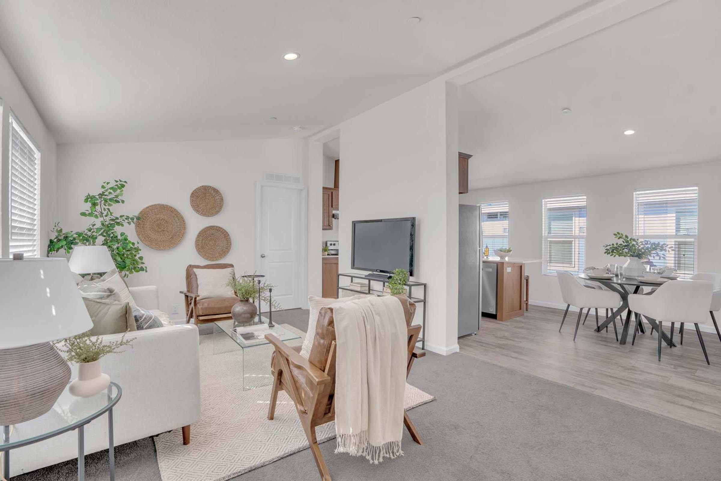 A modern, bright living room features a cozy seating area with a white sofa and wooden chairs. Decorative plants and wall art enhance the space. A television stand and dining area with light-colored furniture are visible in the background, creating a light and inviting atmosphere.