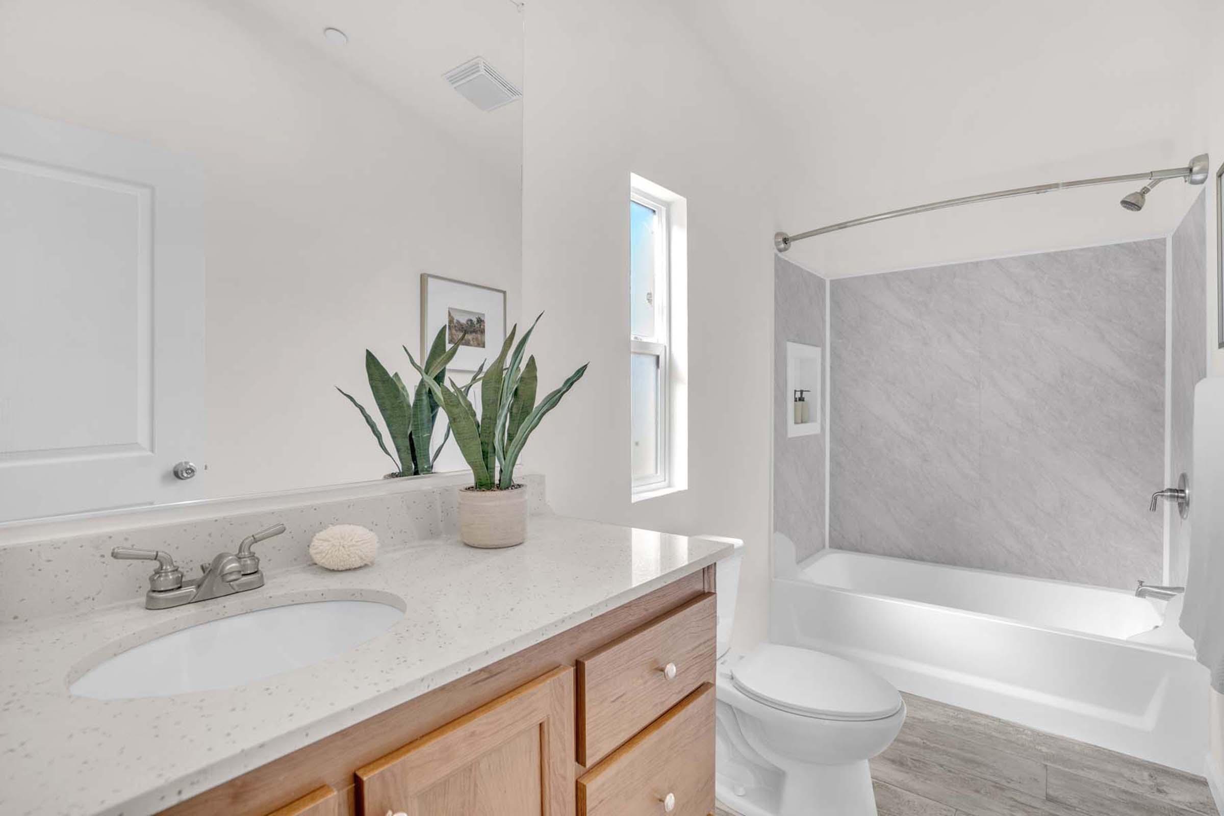 A modern bathroom featuring a clean white aesthetic, equipped with a bathtub and shower combination, a stylish countertop sink with wooden cabinets, and a potted plant for decoration. Natural light enters through a small window, enhancing the bright and airy feel.