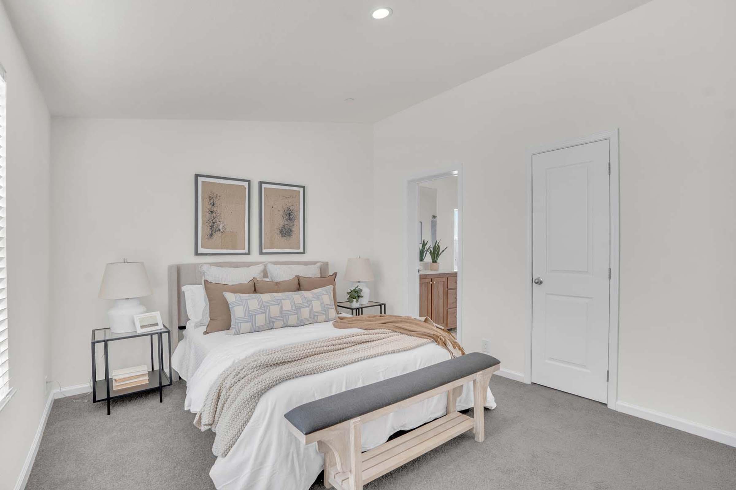 A bright and airy bedroom featuring a queen-sized bed with decorative pillows and a beige throw blanket. There are two framed artwork pieces on the wall, bedside lamps, and a wooden bench at the foot of the bed. A doorway leads to a bathroom, with neutral-colored walls and carpet.