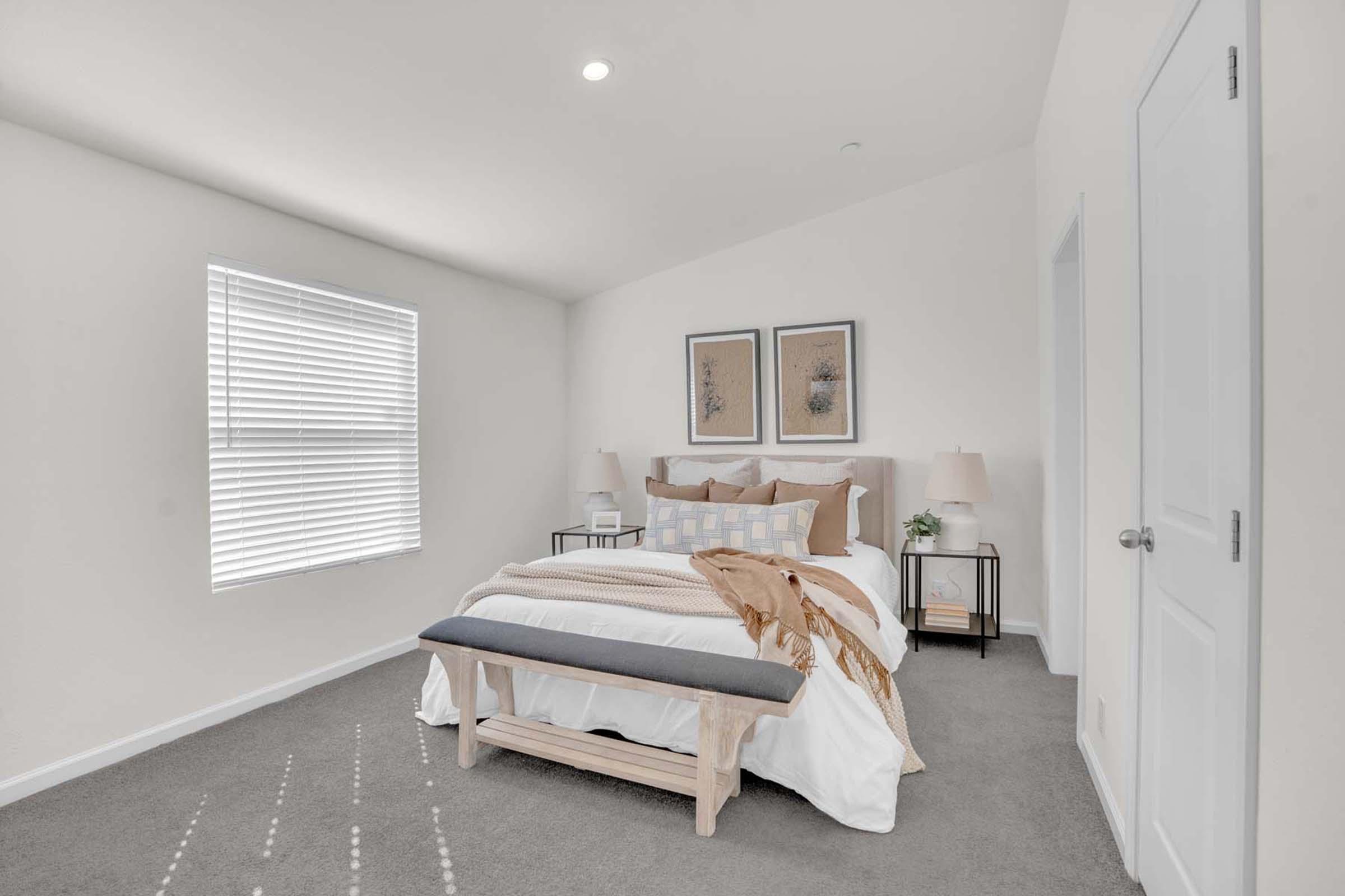 A bright, modern bedroom featuring a neatly made bed with decorative pillows and a soft throw blanket. There are two bedside tables with lamps, a wooden bench at the foot of the bed, and framed artwork on the walls. Natural light streams through a window with blinds. The space has a clean, minimalist design.