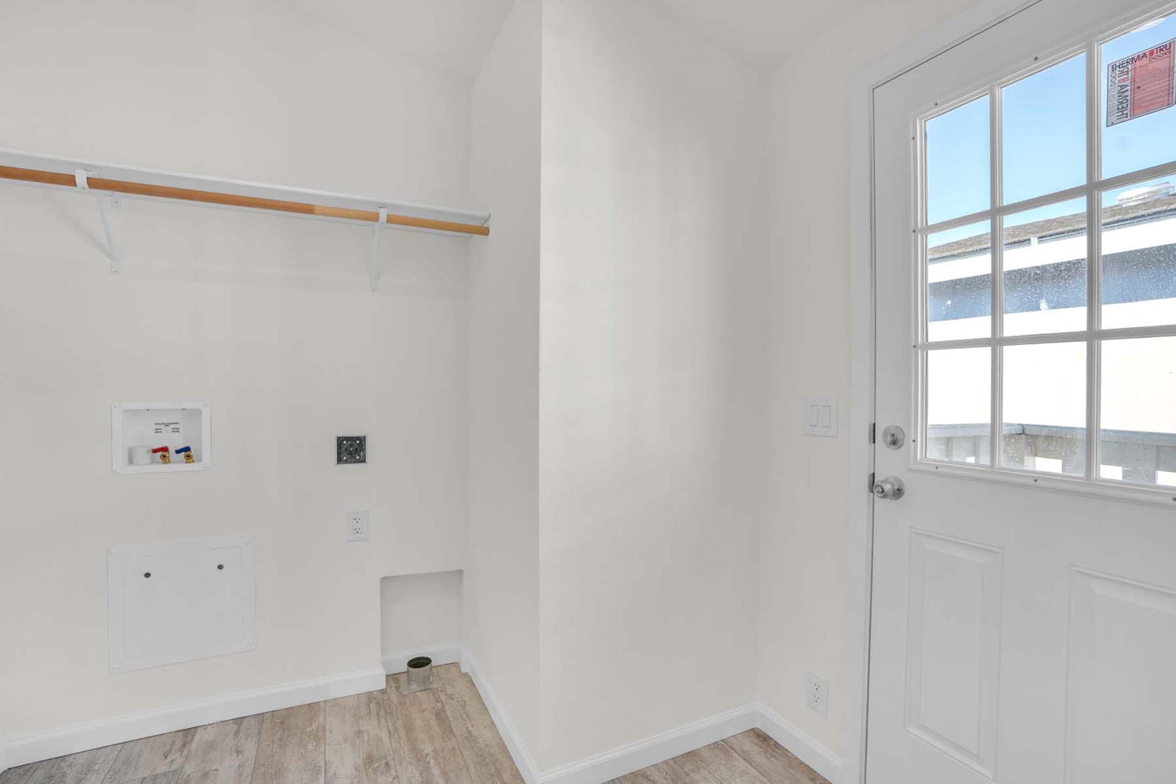 A bright, empty laundry room featuring a small utility shelf, a wall outlet, and a door with multiple square windows leading outside. The walls are painted white, and the flooring is light-colored.