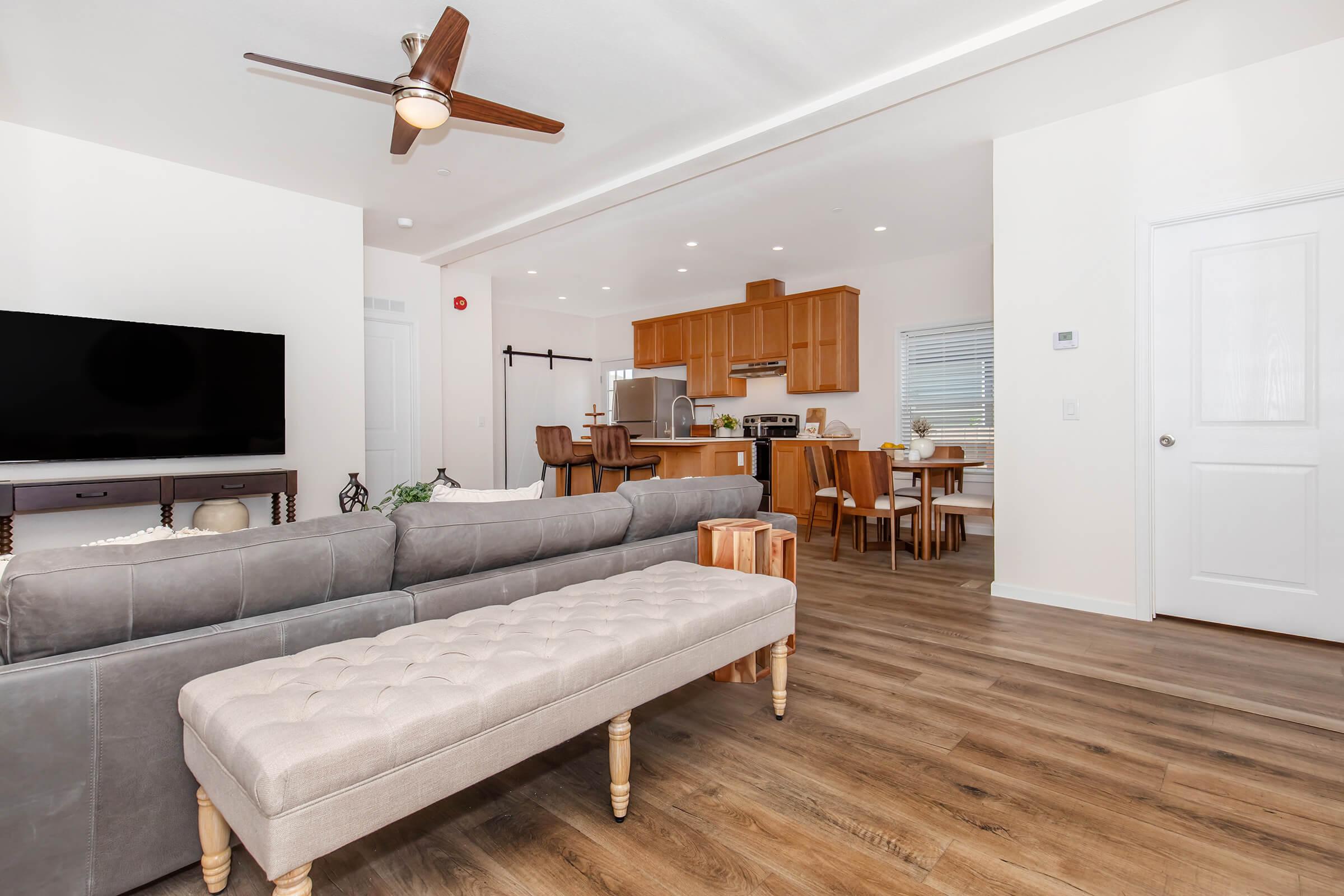 A modern living room featuring a gray sofa with a tufted ottoman, a large flat-screen TV on a wooden console, and a light-filled dining area in the background. The kitchen includes wooden cabinets and appliances. The space has warm wood flooring and bright, neutral-colored walls, creating a cozy atmosphere.