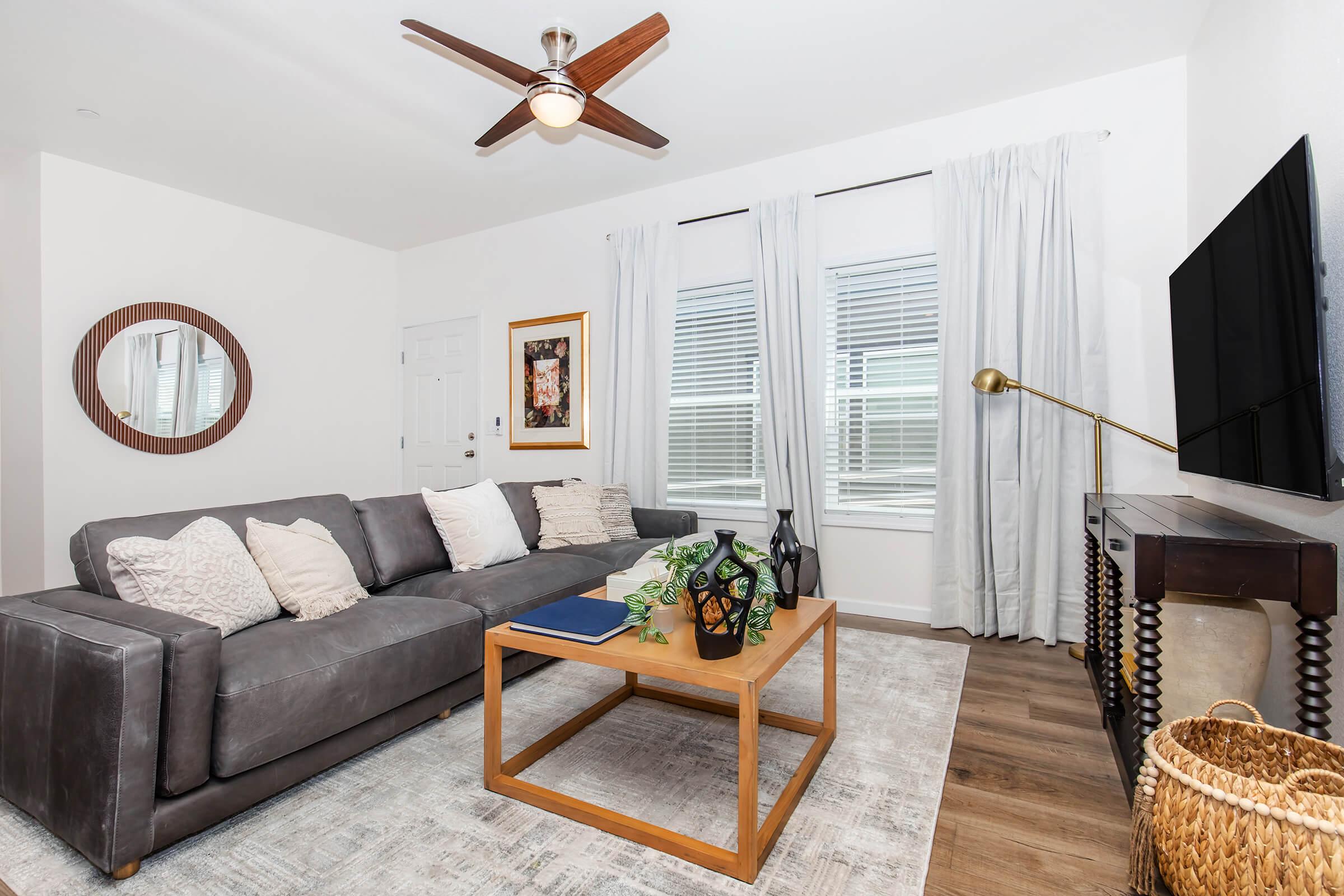 A modern living room featuring a gray couch with decorative pillows, a coffee table with books and a decorative vase, and a wall-mounted television. Natural light streams through large windows, complemented by sheer white curtains. A round mirror hangs on the wall, and a cozy woven basket adds texture to the space.