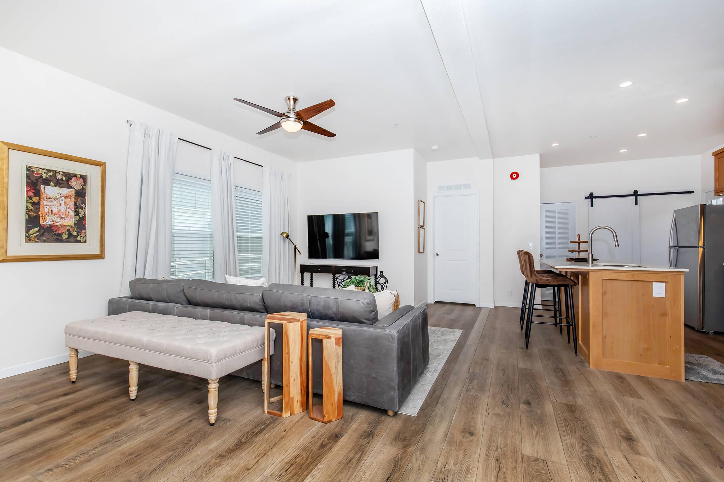 A modern living space featuring a gray sofa with a beige ottoman, a wooden coffee table, and a flat-screen TV mounted on the wall. Natural light filters through sheer curtains, illuminating the room. In the background, there’s a kitchen area with wooden cabinetry and bar stools.