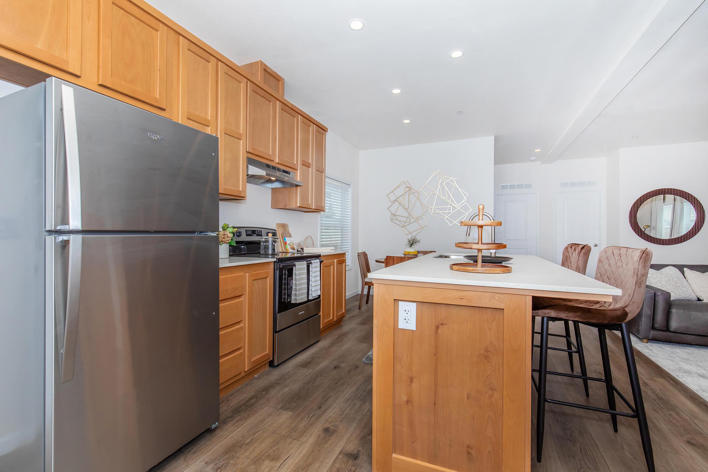 Modern kitchen featuring stainless steel appliances, including a refrigerator and oven. Warm wood cabinetry complements a spacious island with seating. Bright, open layout with natural light, and a cozy living area visible in the background. Elegant decor and contemporary design elements throughout.