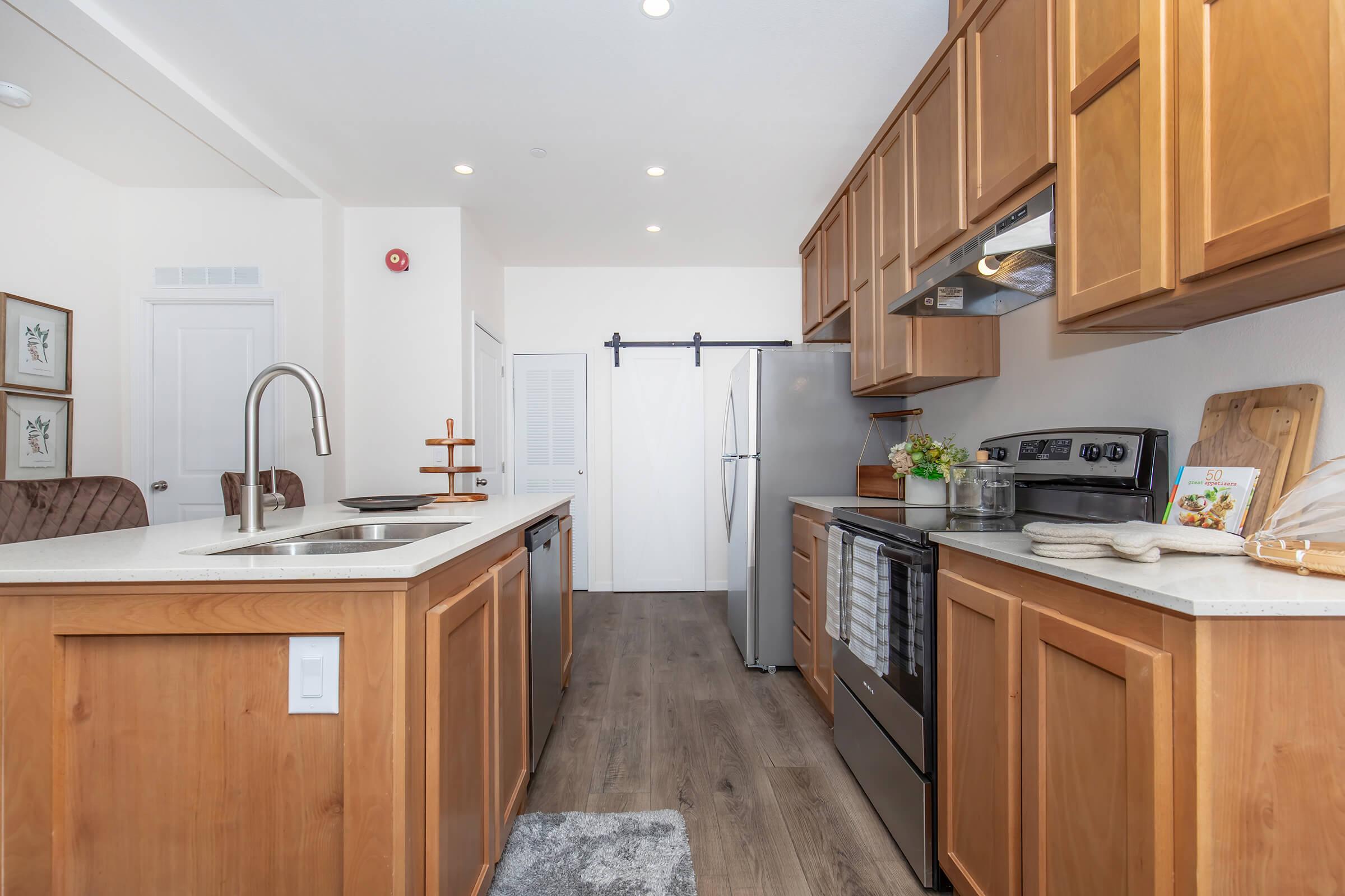 Modern kitchen featuring wooden cabinetry, stainless steel appliances, and a central island with a sink. Brightly lit with overhead lights, the space has a sleek design and includes a refrigerator, stove, and decorative elements like plants and cookbooks, creating a warm and inviting atmosphere.