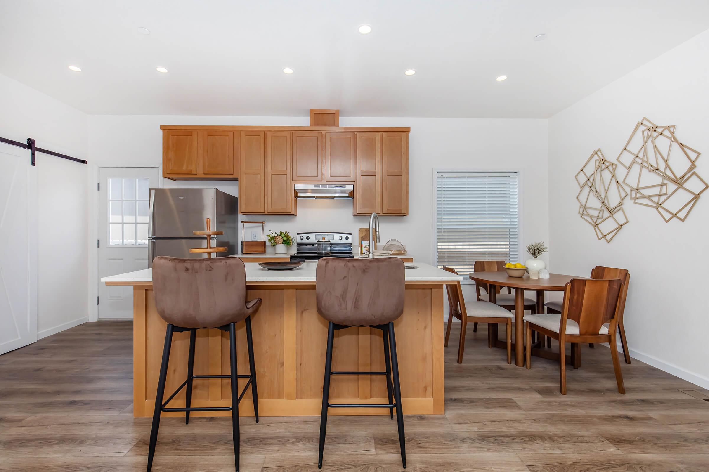 Modern kitchen featuring wooden cabinets and stainless steel appliances. A central island with bar stools, and a dining table set for four. Natural light fills the space through the window, complemented by decorative wall art in geometric shapes. The flooring is a warm wood tone, creating a cozy atmosphere.