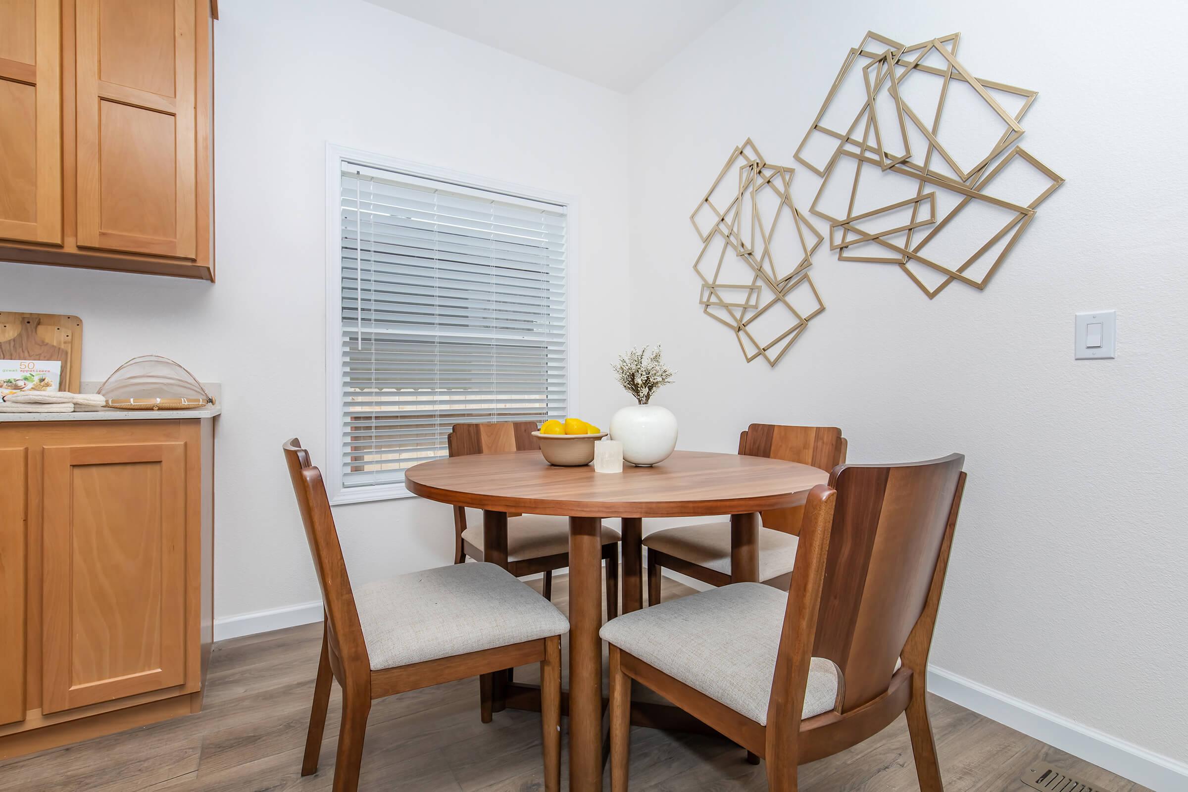 A cozy dining area featuring a round wooden table surrounded by four wooden chairs with light-colored cushions. A bowl of lemons sits in the center of the table, and a small vase with dried flowers adds a touch of decor. The background includes a window covered with white blinds and decorative wall art in geometric shapes.