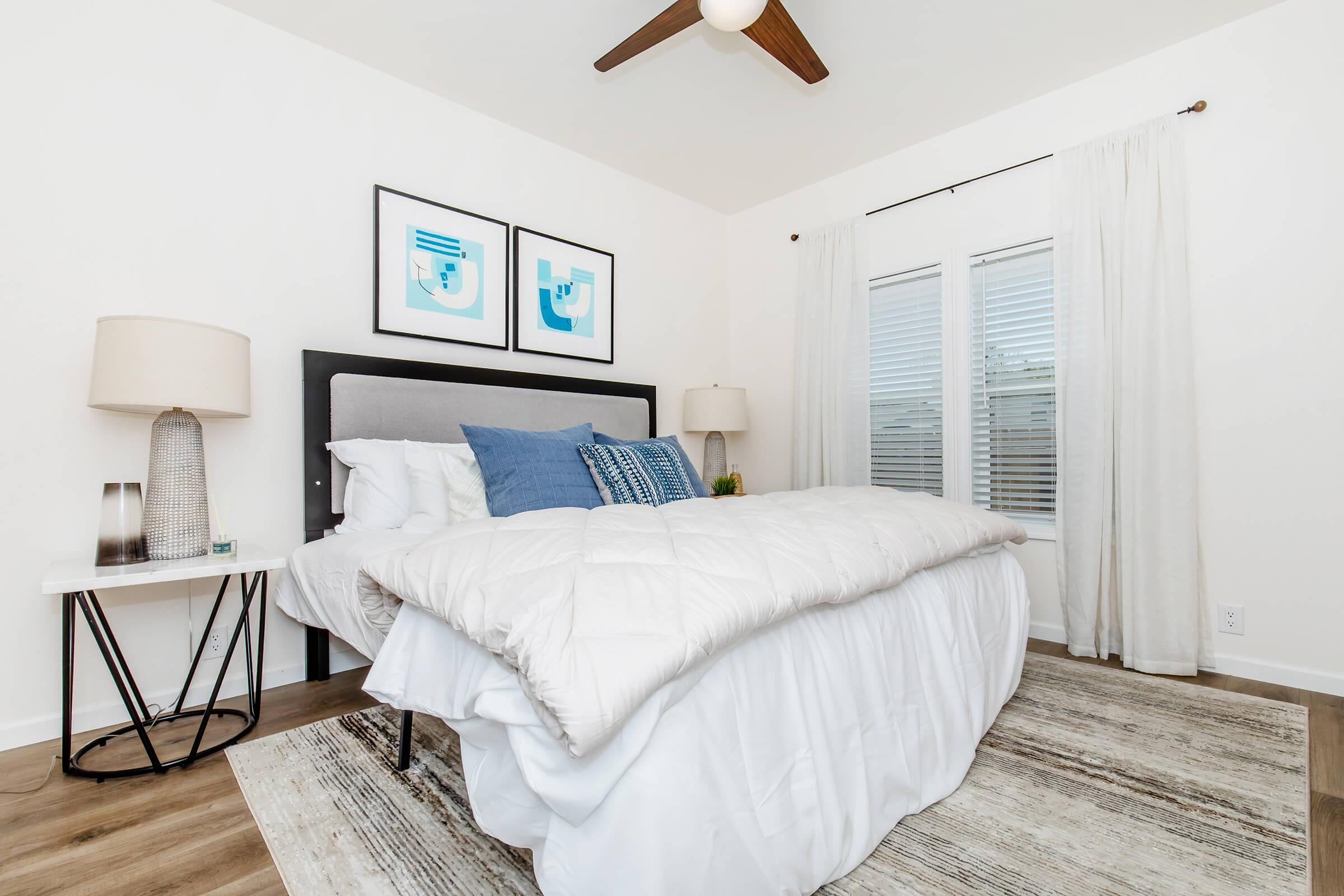 A modern bedroom featuring a neatly made bed with a white comforter and blue accent pillows. Two framed abstract artworks hang on the wall above the bed. There are bedside tables with lamps on either side, and a window with sheer curtains lets in natural light, illuminating the light wooden floor.