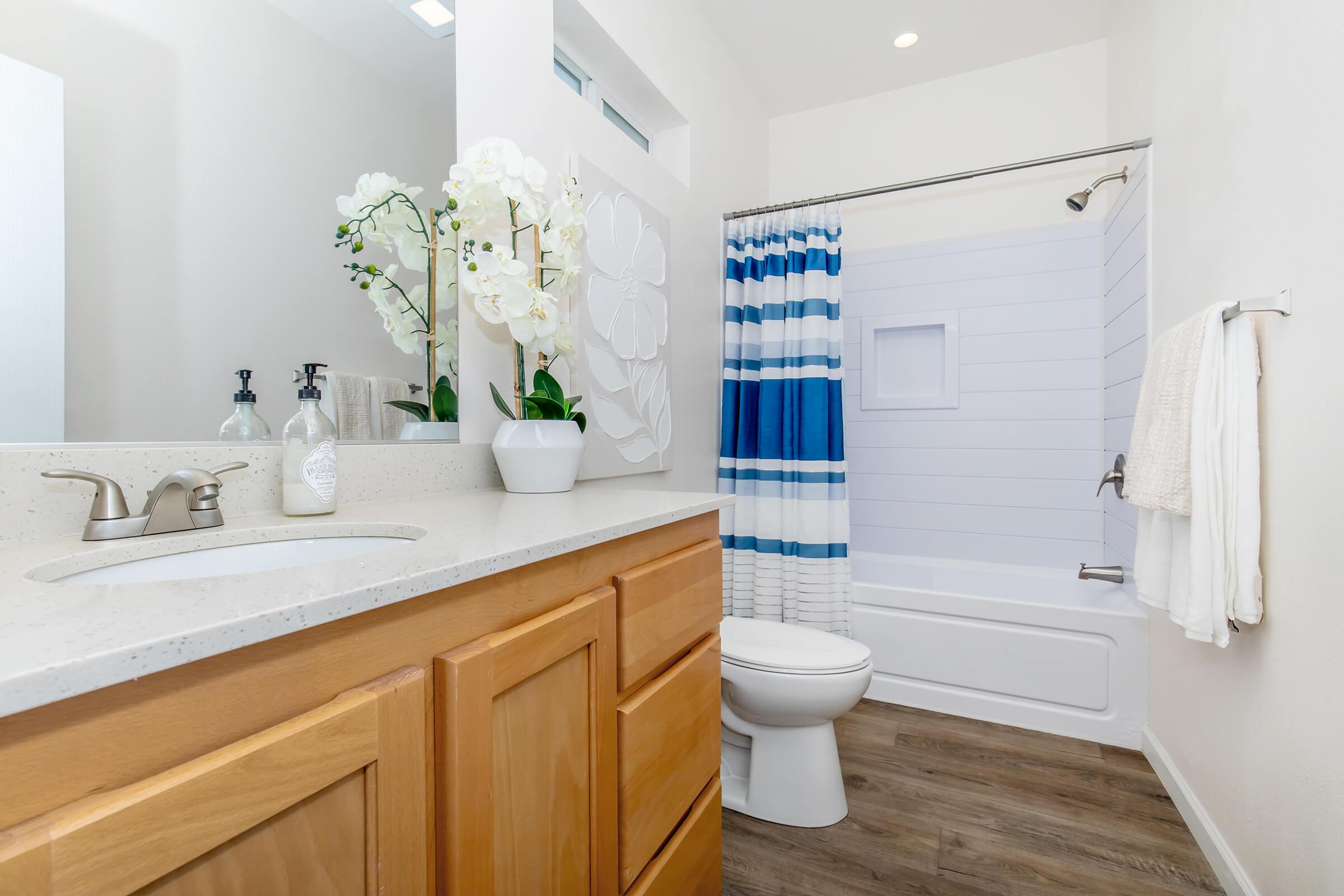 A modern bathroom featuring a light wooden vanity with a sink, a white toilet, and a shower area with a blue and white striped curtain. Decorative elements include a potted orchid and stylish hand soaps. The space has neutral-colored walls and a clean, bright atmosphere.
