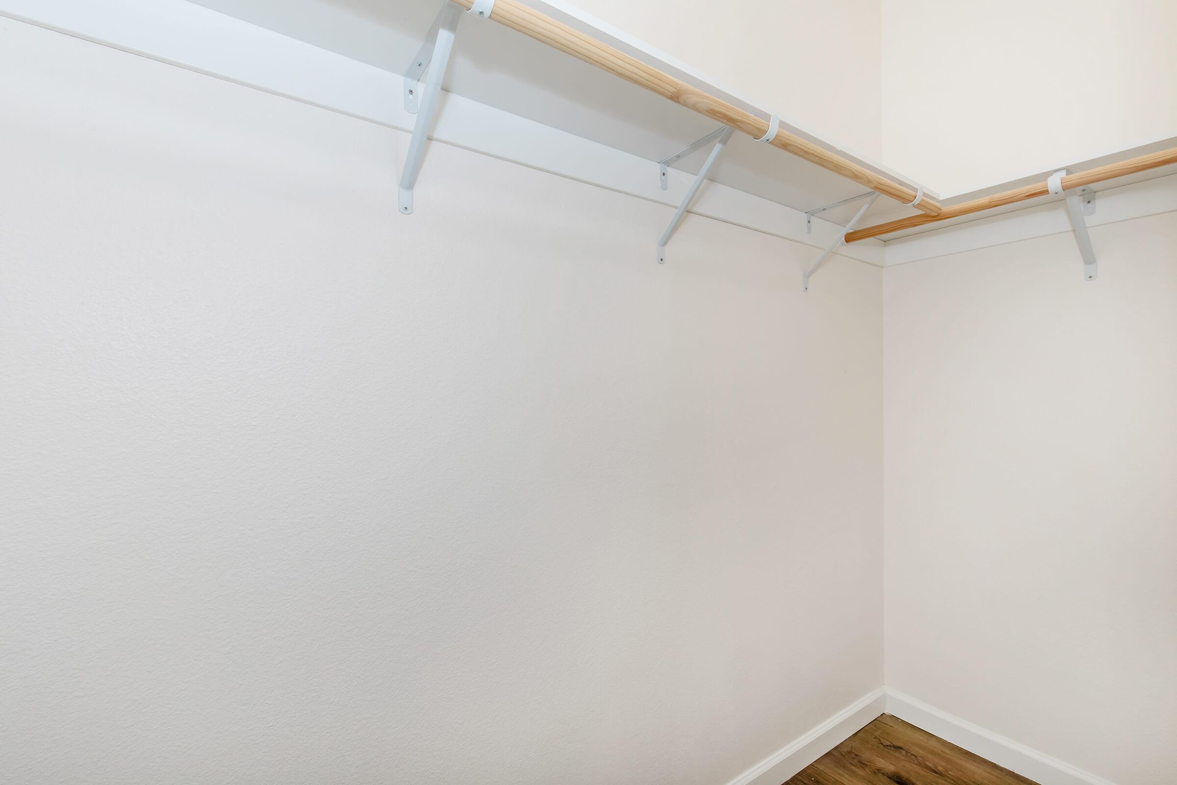 Empty closet with white walls and a wooden shelf mounted on a white support system. The floor has a light wooden finish, and there is ample natural light illuminating the space.