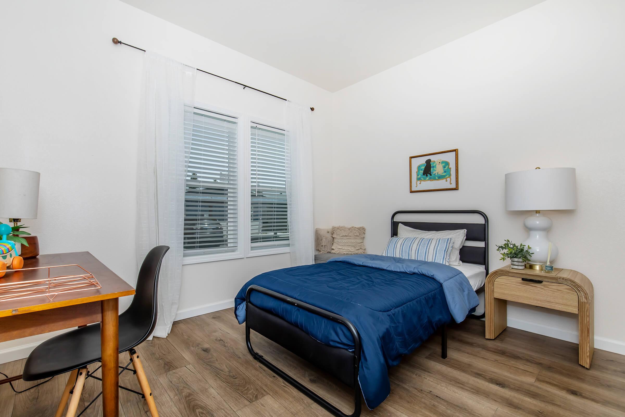 A cozy bedroom featuring a single bed with a blue duvet, a desk with a wooden top and a black chair, a nightstand with a table lamp, and a framed picture on the wall. Natural light streams in through a window adorned with white curtains, illuminating the warm wood flooring and minimalistic decor.