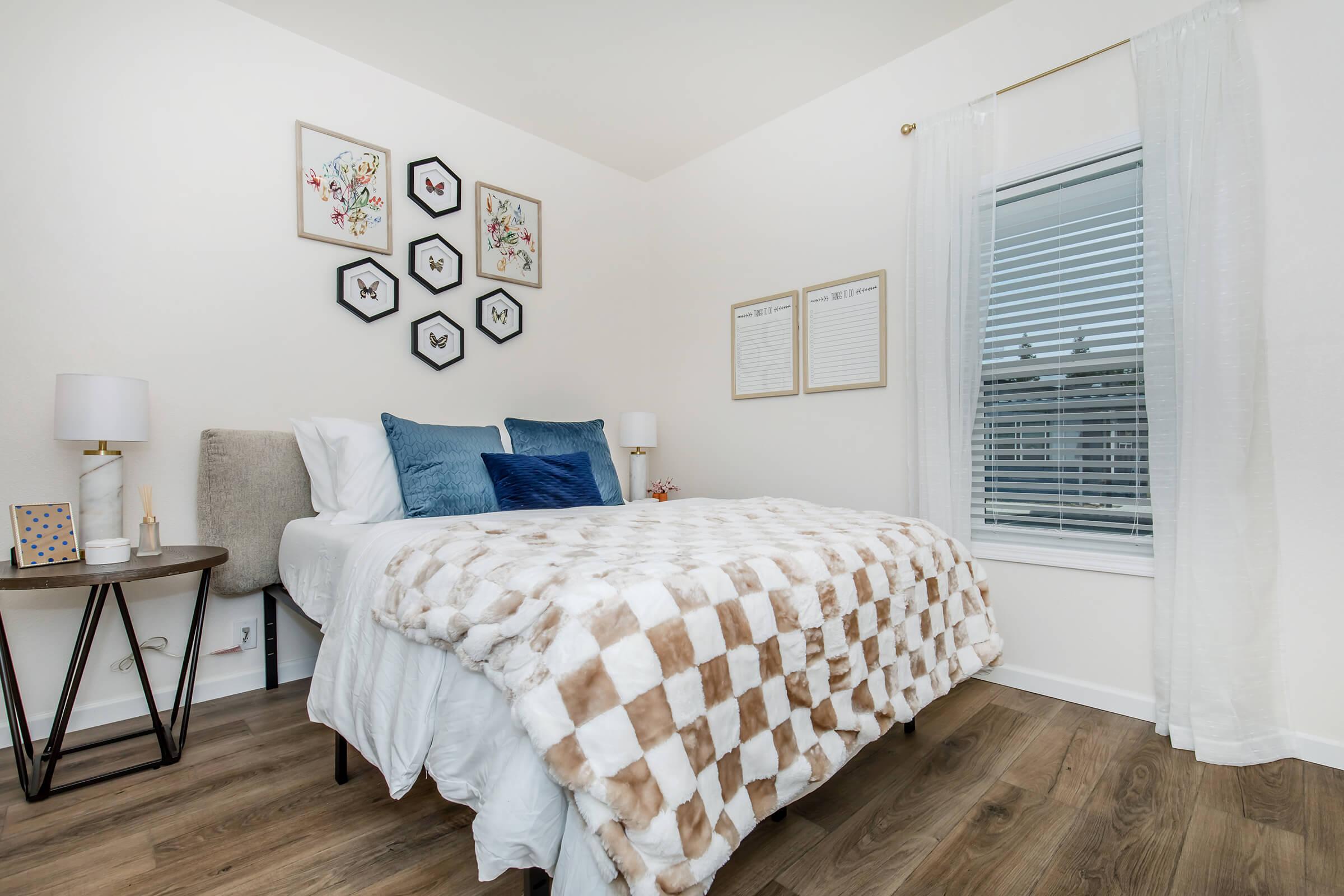 A cozy bedroom featuring a neatly made bed with a fluffy checkered blanket, blue and white decorative pillows, and framed butterfly artwork on the wall. A wooden nightstand with a lamp and decorative items stands next to the bed, and a window with sheer curtains allows natural light to brighten the space.