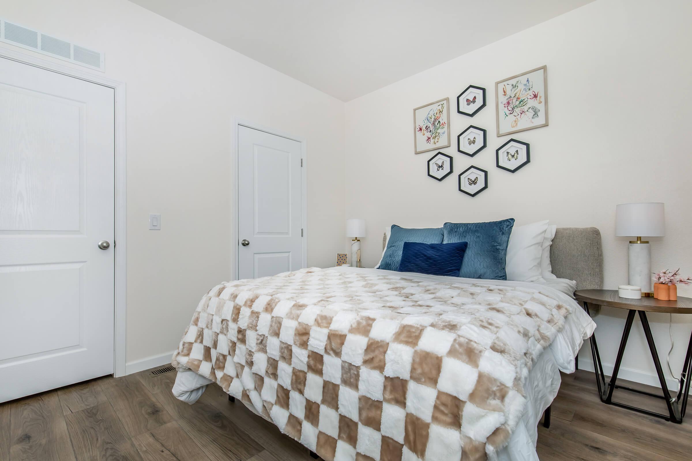 A cozy bedroom featuring a neatly made bed with a beige and white checkered blanket, blue and white pillows. A light-colored wall displays a decorative arrangement of framed butterfly artwork. A bedside table holds a lamp, and a closed door is visible in the background, adding to the inviting atmosphere.