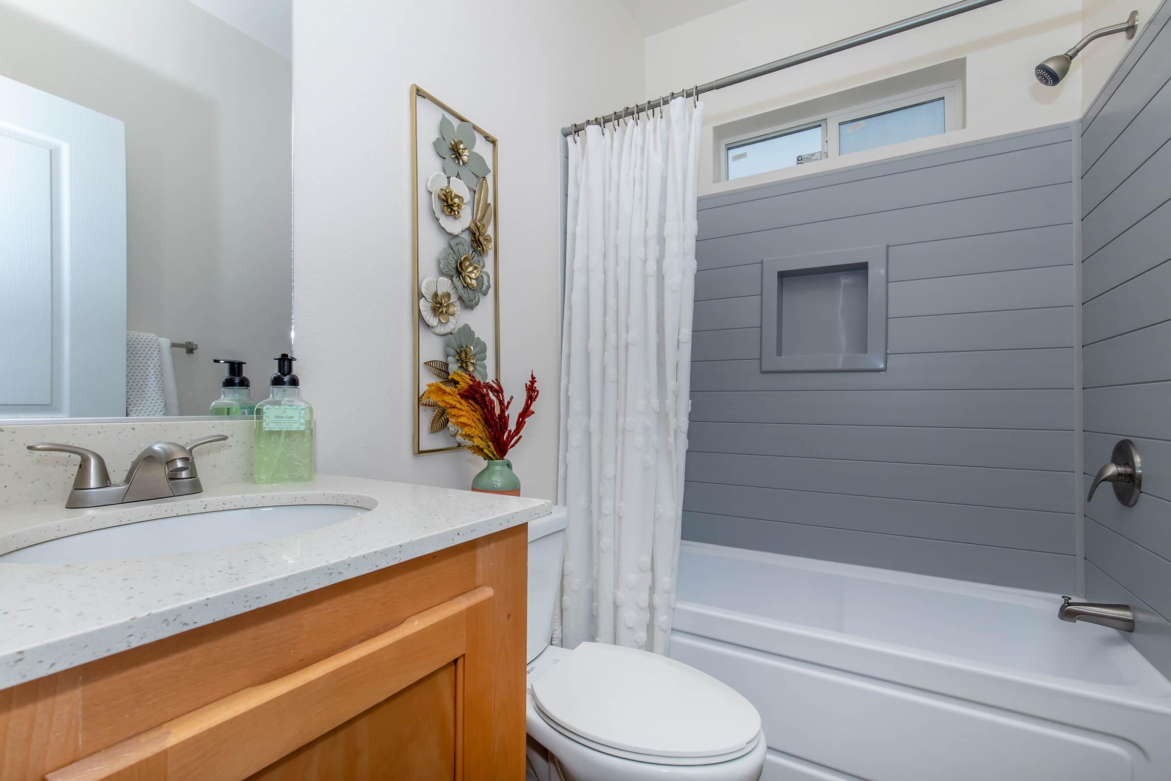 A modern bathroom featuring a bathtub with a gray panel, a shower curtain, and a light-colored countertop. Decorative wall art with flowers is mounted beside the bathtub. There are toiletries on the counter and a mirror reflecting part of the space. The overall aesthetic is clean and minimalist.