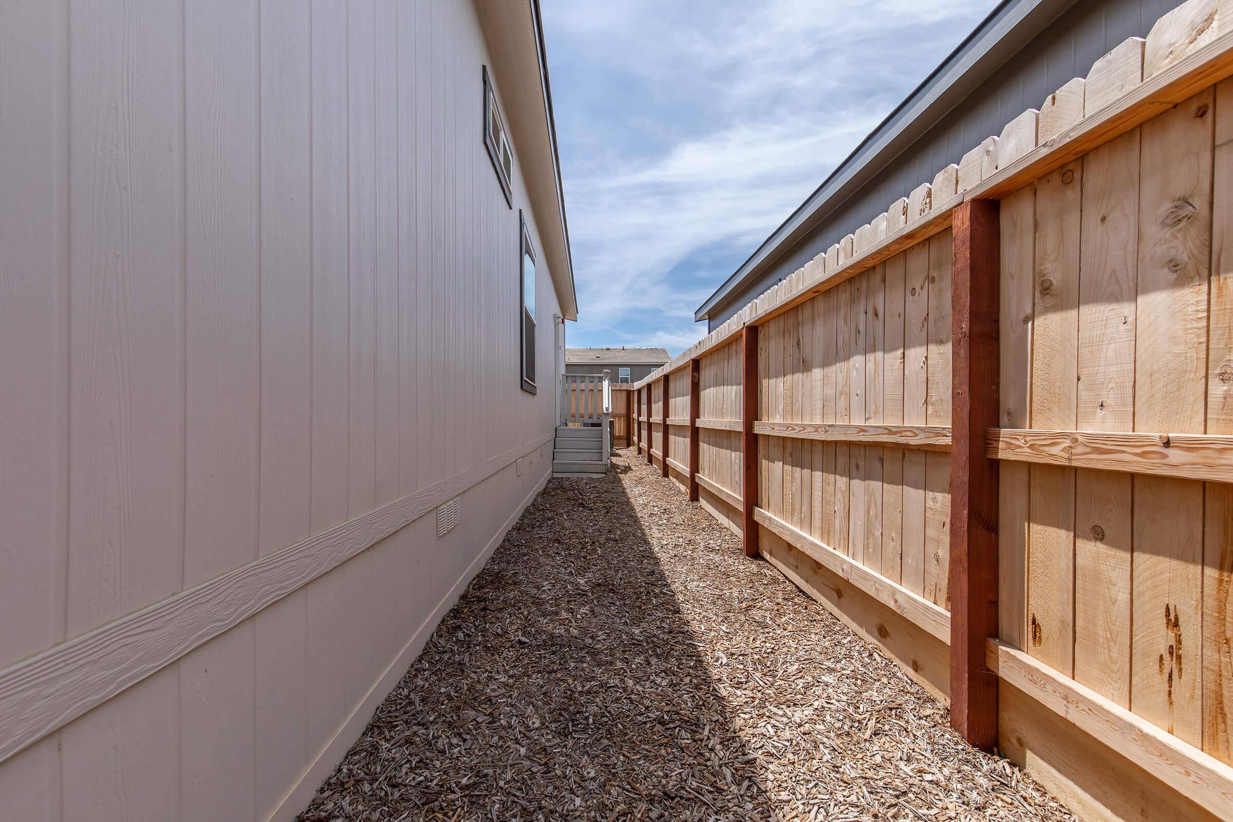 Narrow pathway between two houses, lined with wooden fences on either side. The ground is covered with wood chips, and the sky is partly cloudy, creating a bright atmosphere.