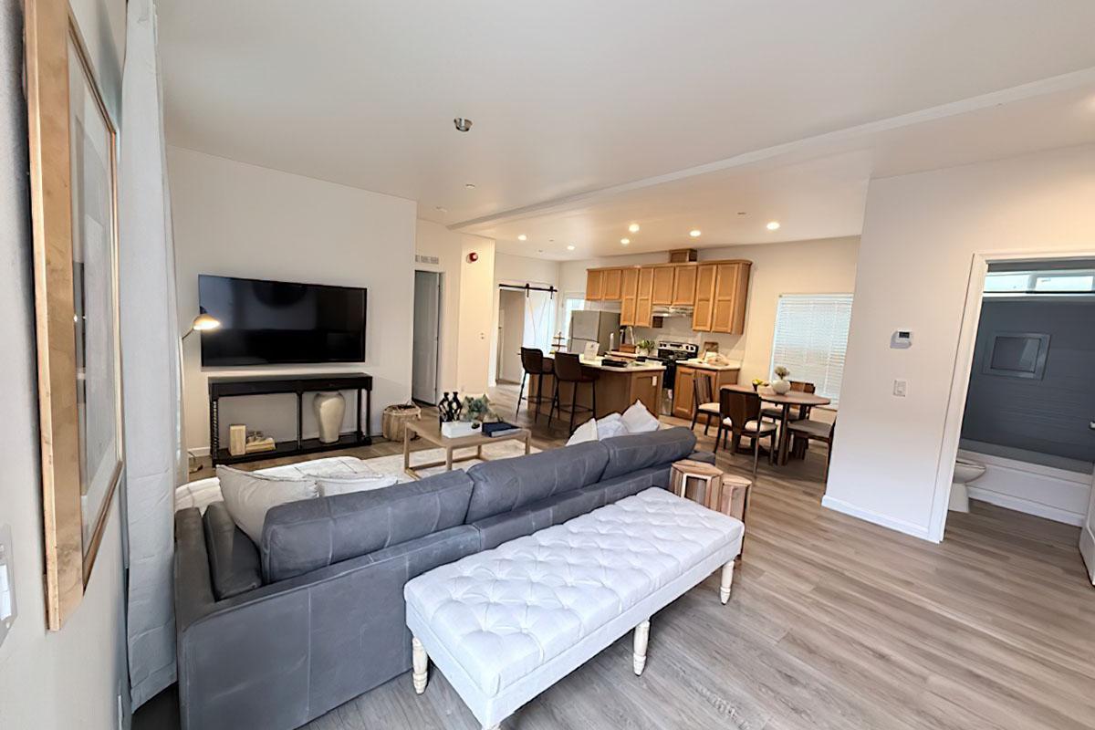 A modern living space featuring a gray sectional sofa, a large TV mounted on the wall, and a wooden dining table with chairs. The open layout connects the living area to a kitchen with wooden cabinets. Natural light enters through windows, creating a bright and inviting atmosphere.