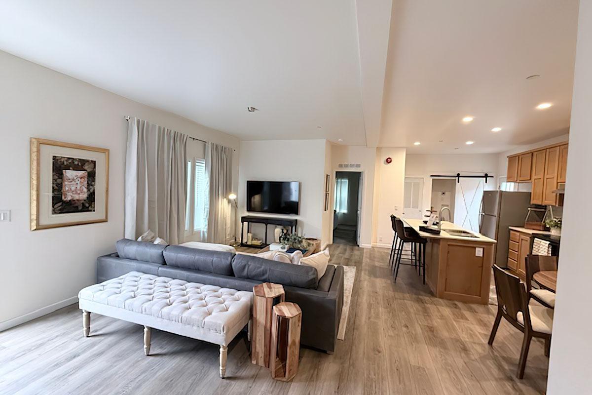 A modern, open-concept living room and kitchen area. The living room features a gray sectional sofa, a framed artwork on the wall, and a flat-screen TV. The kitchen is visible in the background, with wooden cabinetry, stainless steel appliances, and a dining area with bar stools. Enjoys natural light from window.