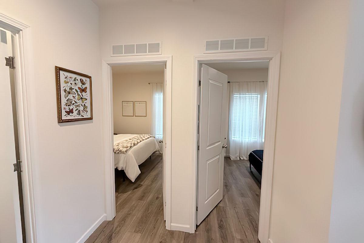 A view of a bright hallway with two doorways leading to separate rooms. One door opens to a room with a bed, while the other leads to a darker room with a couch. Natural light filters in through windows, and there is a framed butterfly artwork on the wall near the entrance. The flooring is light-colored wood.