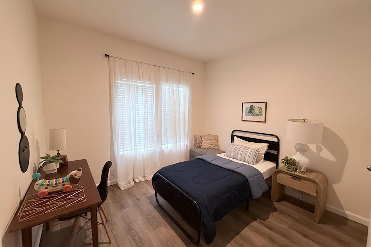 A cozy bedroom featuring a black metal bed with a blue blanket, a wooden desk with a colorful decorative item, a comfortable chair, a large lamp, and a small side table with a plant. Natural light filters through sheer white curtains over a window, creating a warm and inviting atmosphere.