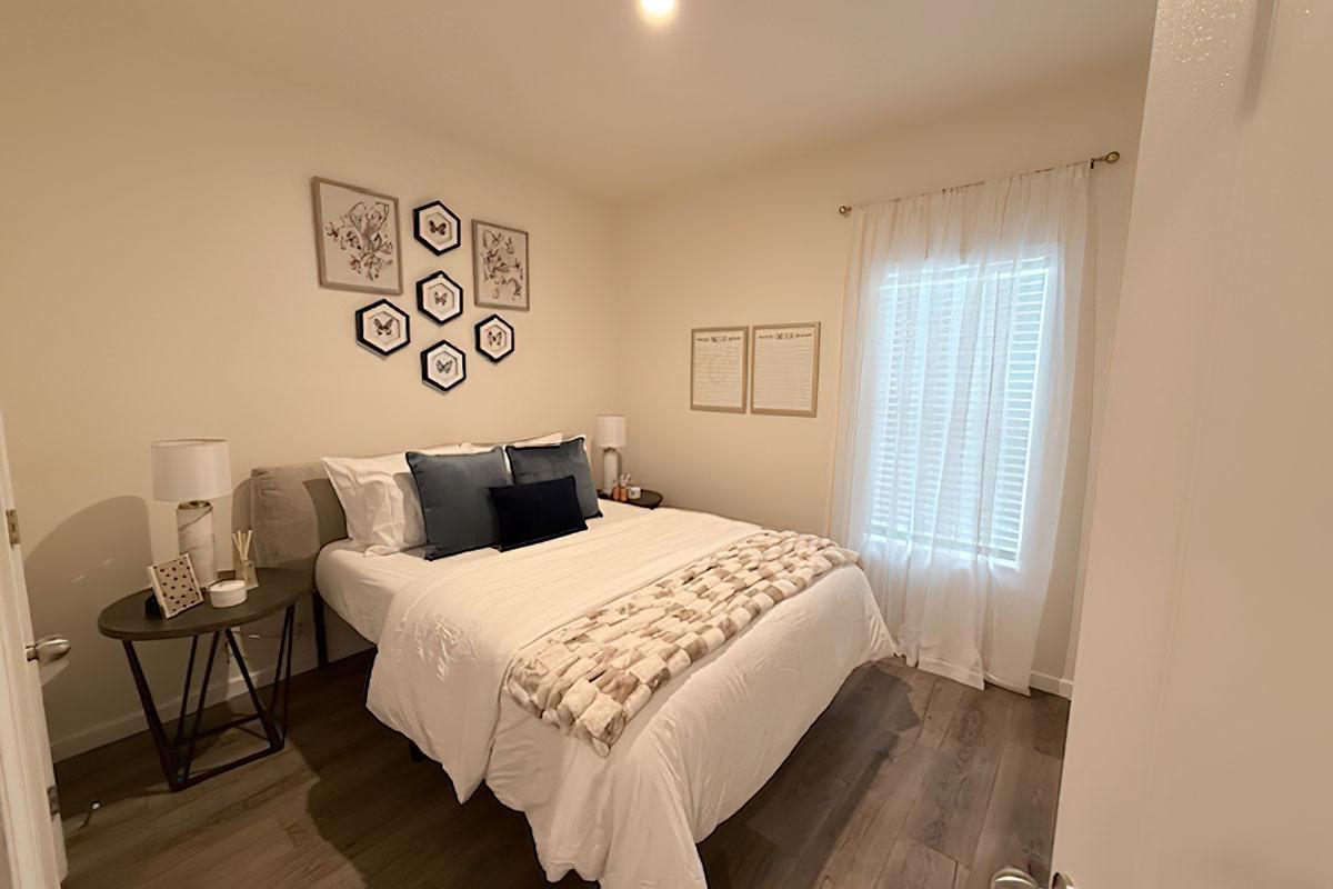 Cozy bedroom featuring a queen-size bed with white bedding and decorative pillows. A small side table holds a lamp. The walls are adorned with framed artwork. Large window with sheer curtains allows natural light, and wooden flooring adds warmth to the space.