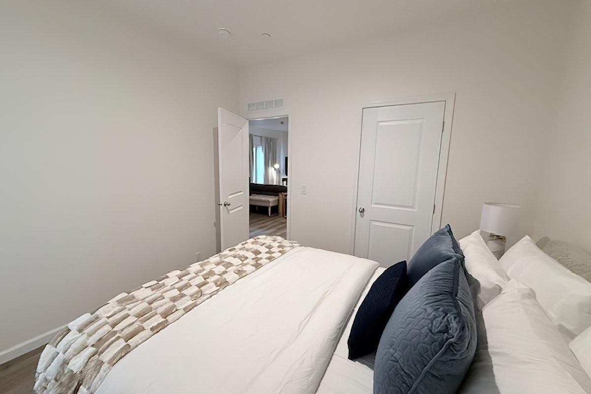 A cozy, minimalist bedroom featuring a neatly made bed with light-colored linens and decorative pillows. The room has soft lighting and an open door leading to another area, emphasizing a clean and inviting atmosphere. Smooth walls and simple decor create a tranquil ambiance.