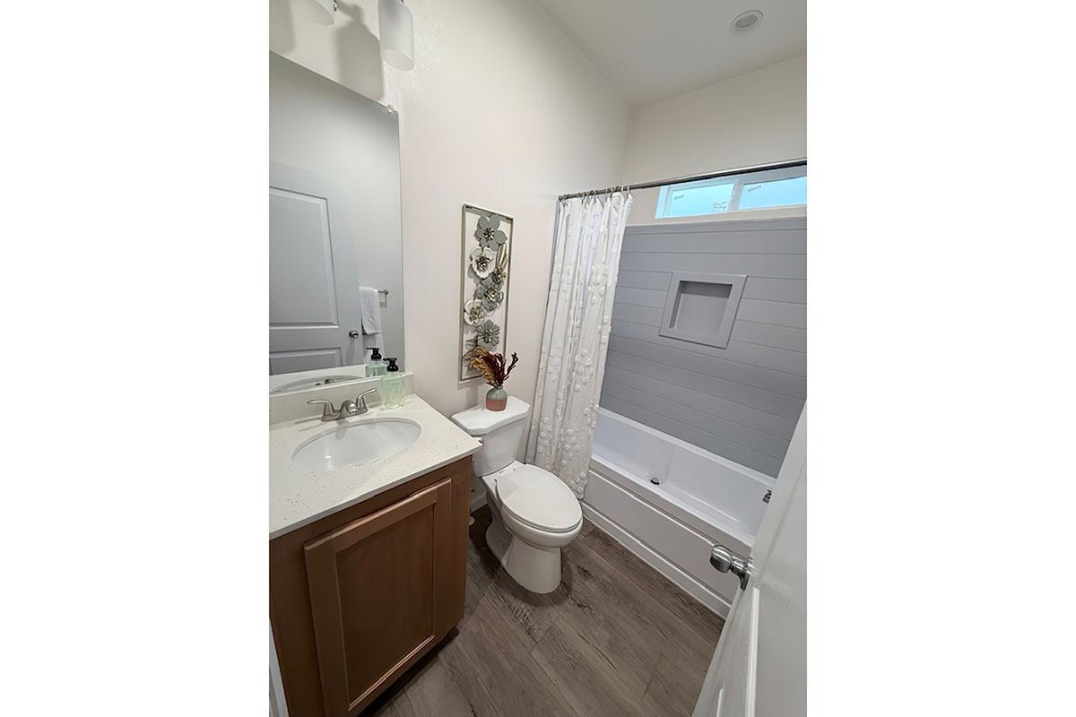 A clean and modern bathroom featuring a white bathtub with a shower curtain, a wooden vanity with a sink, a mirror above the sink, and decorative wall art. The floor has light-colored tiles, and there is a small window allowing natural light. The space is well-organized and bright.