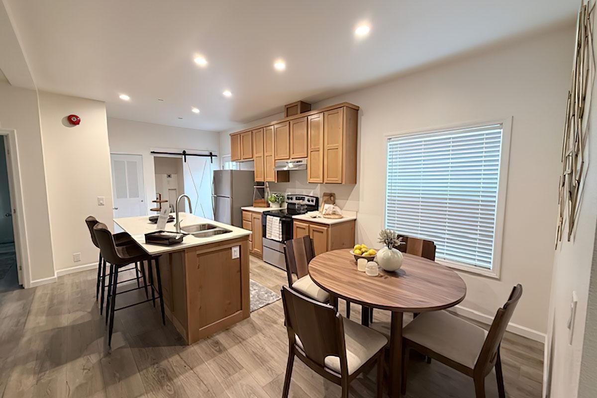 A modern kitchen featuring wooden cabinets, stainless steel appliances, and a small dining area with a round table and chairs. The space is well-lit with recessed lighting, and there is a window with blinds letting in natural light. A fruit bowl adds a touch of decor on the table.