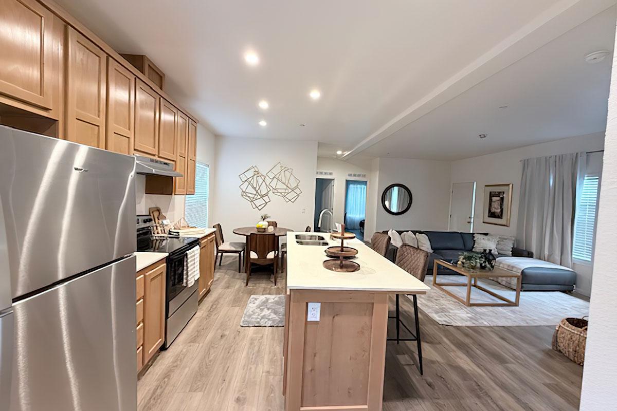 Modern kitchen and living room interior featuring stainless steel appliances, wooden cabinets, and a large island with seating. The open layout includes a dining area, a cozy sofa, and decorative wall art. Natural light streams in through windows, creating a warm and inviting atmosphere.