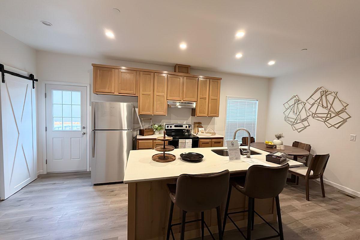 Modern kitchen featuring wooden cabinets, stainless steel appliances, and a large island with seating. A dining table is positioned nearby, and there are decorative wall accents. Brightly lit with natural light coming through a window and a door leading outside. Stylish and functional space for cooking and dining.