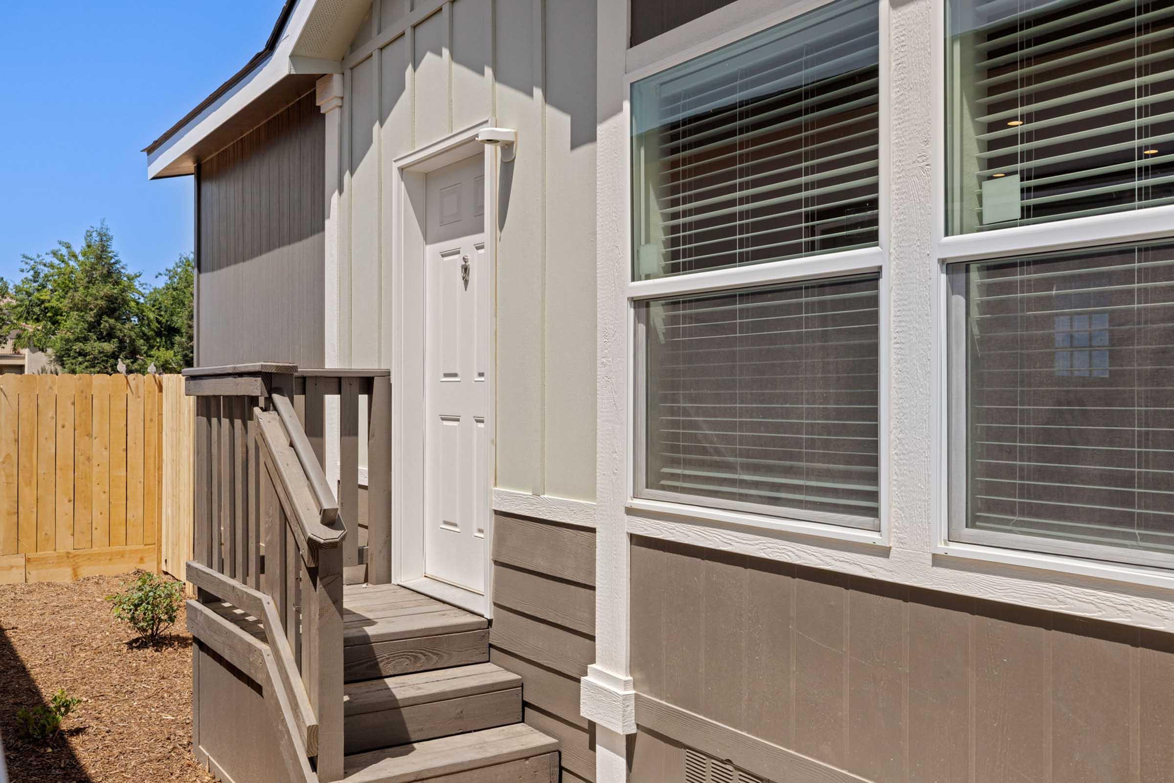 Exterior view of a house showing a staircase leading up to a white door. The house features light gray siding and large windows with blinds. A wooden fence and landscaping are visible in the background under a clear blue sky.