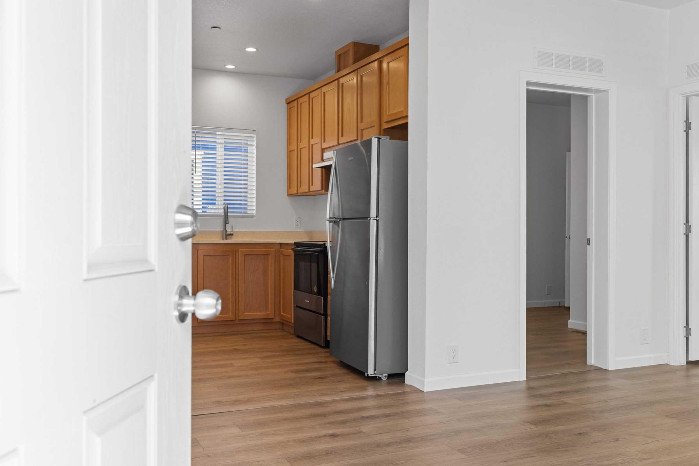 A view of a modern kitchen from an open doorway, featuring stainless steel appliances, wooden cabinets, and a sleek countertop. The room has a clean and bright atmosphere with natural light coming through a window. The flooring is light-colored wood, creating a spacious and inviting feel.