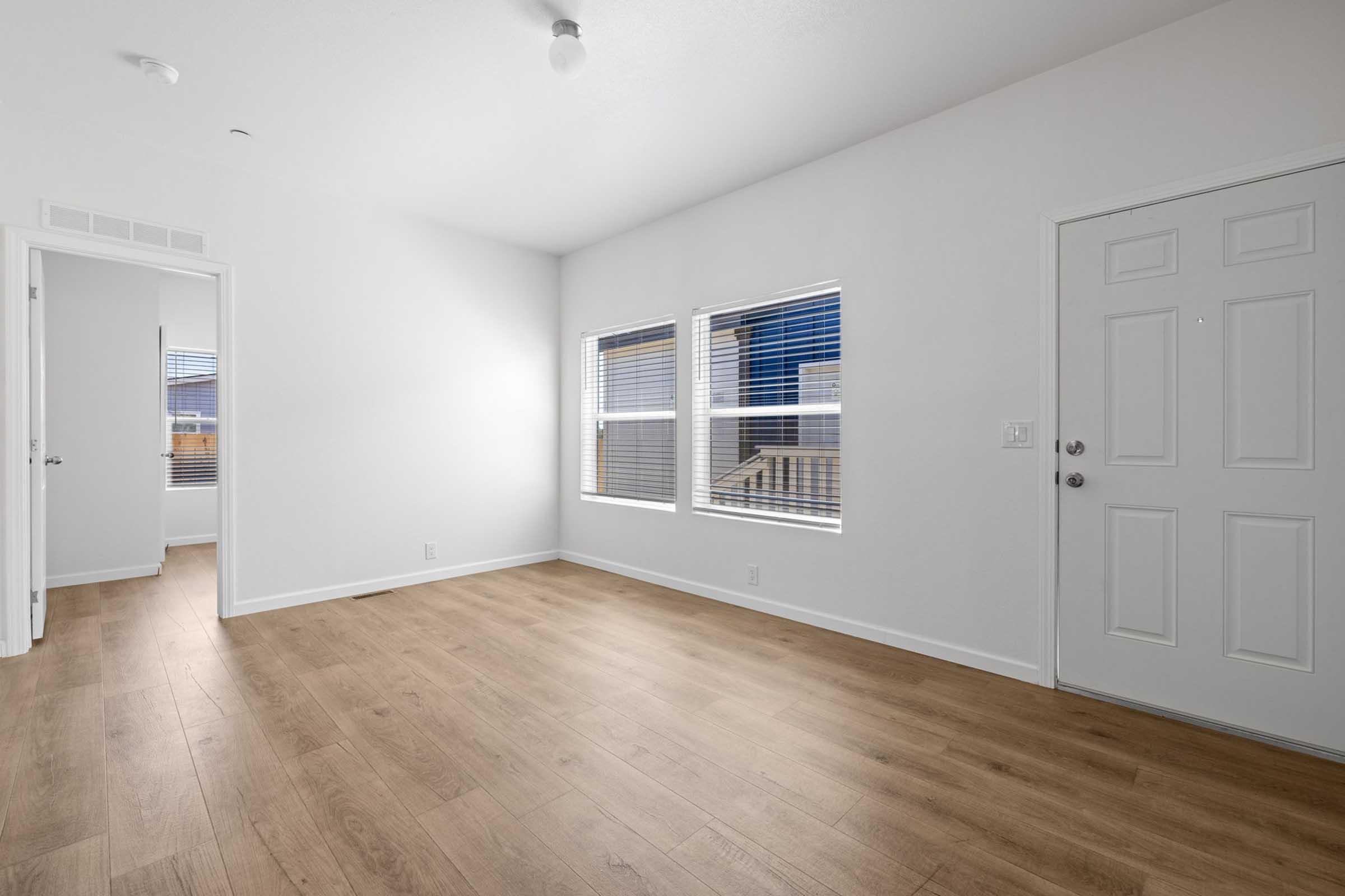 A spacious, empty room with white walls and large windows that allow natural light to enter. The area features light-colored wooden flooring and includes a door leading outside. A hallway is visible in the background, suggesting additional rooms or spaces.