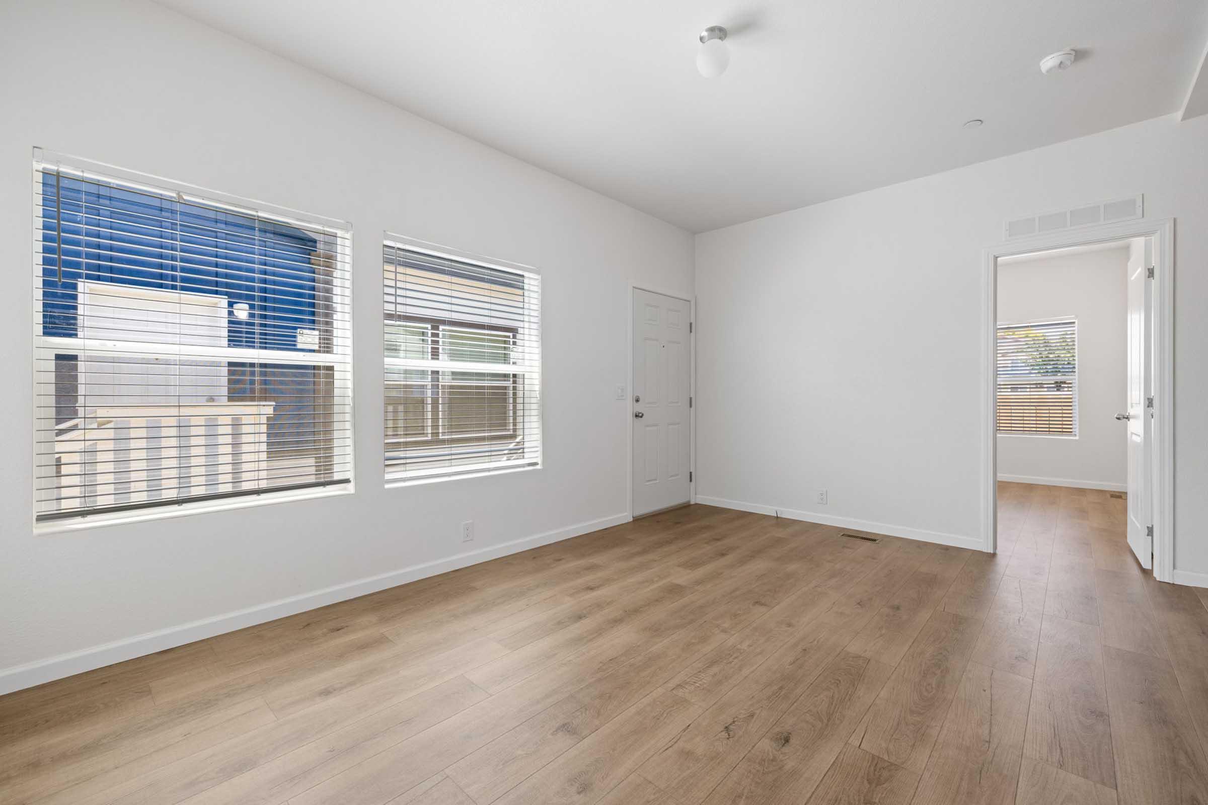 Bright, empty room with wood-style flooring and large windows featuring horizontal blinds. The walls are painted white, and there is a single light fixture on the ceiling. A door leads to another room, and there's a view of an outdoor area through the windows.