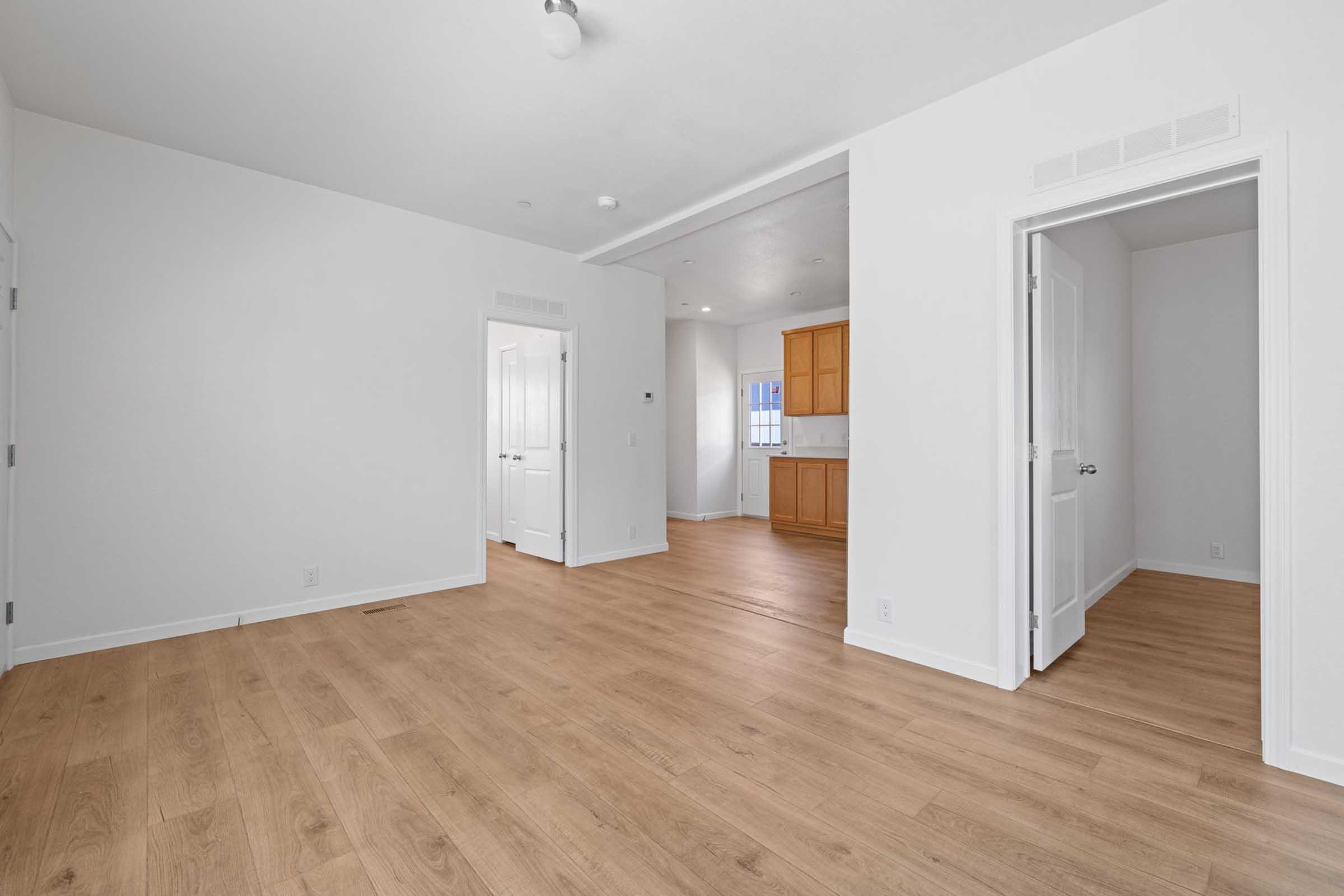 A bright, empty room with light-colored walls and wooden flooring. Two doors are visible leading to adjacent spaces. One door opens to a well-lit kitchen area with wooden cabinets in the background. Natural light floods the room through a window, creating a spacious and inviting atmosphere.