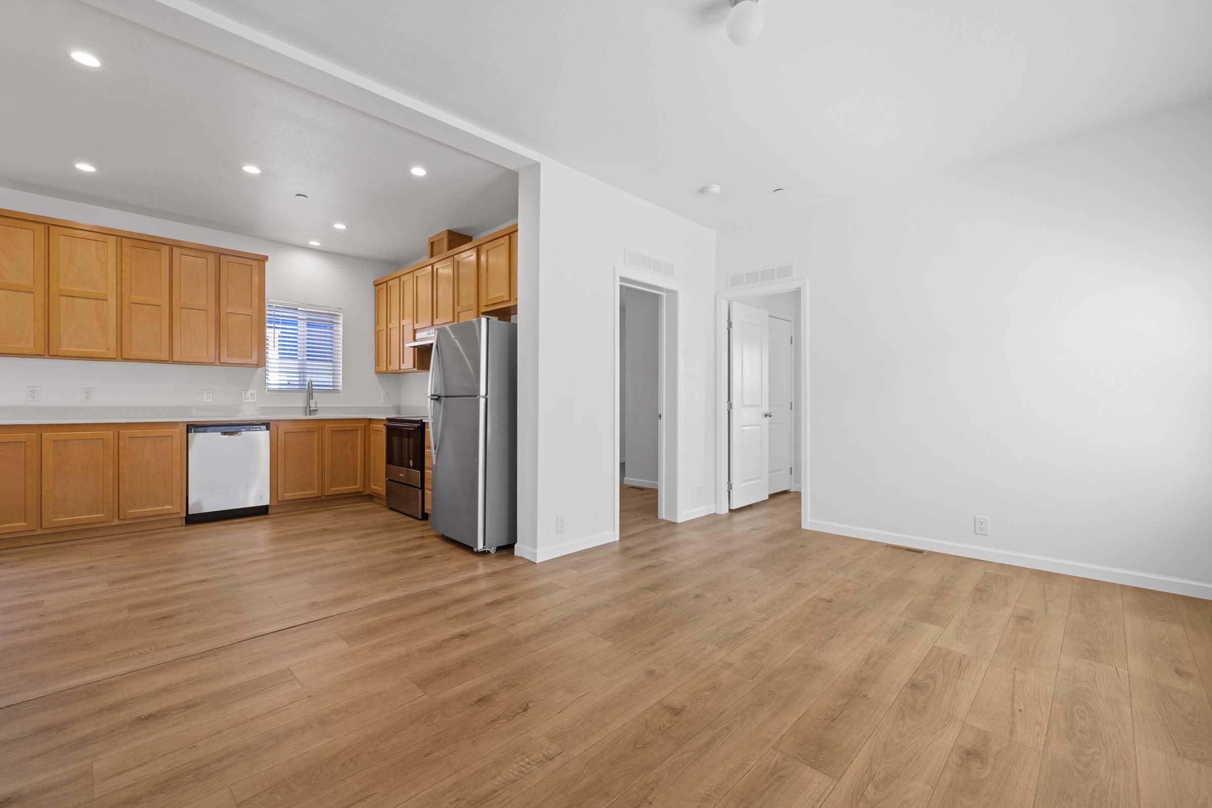 Bright and modern kitchen and living area featuring light wood cabinets, stainless steel appliances, and a spacious layout. The kitchen includes a refrigerator, dishwasher, and window for natural light. The living area has light-colored walls and wooden flooring, creating an open and inviting atmosphere.