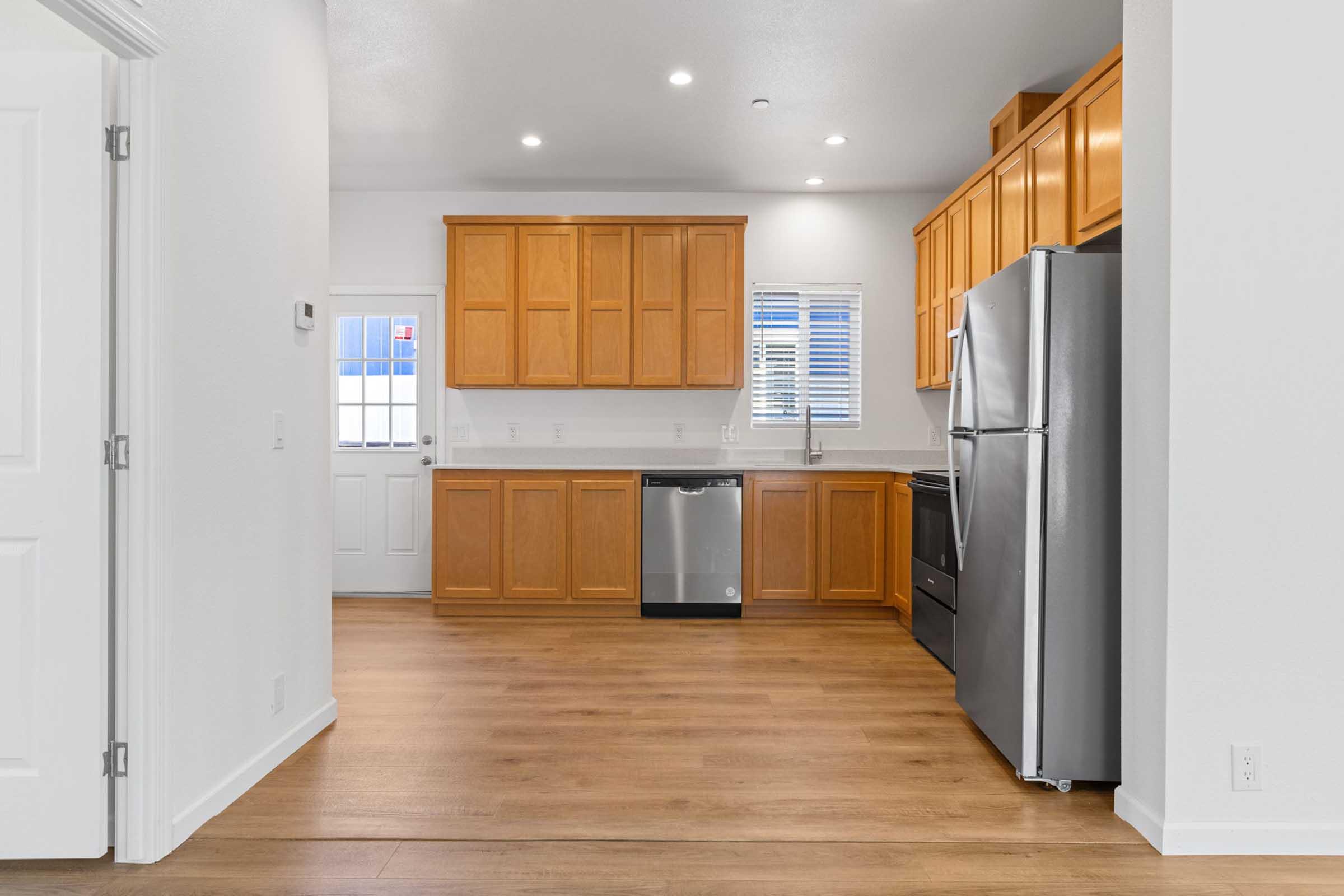 A modern kitchen featuring wooden cabinetry, stainless steel appliances including a refrigerator, dishwasher, and oven. The space has a light-colored countertop, hardwood flooring, and a window with blinds. A door leads to the outside, providing natural light to the area.