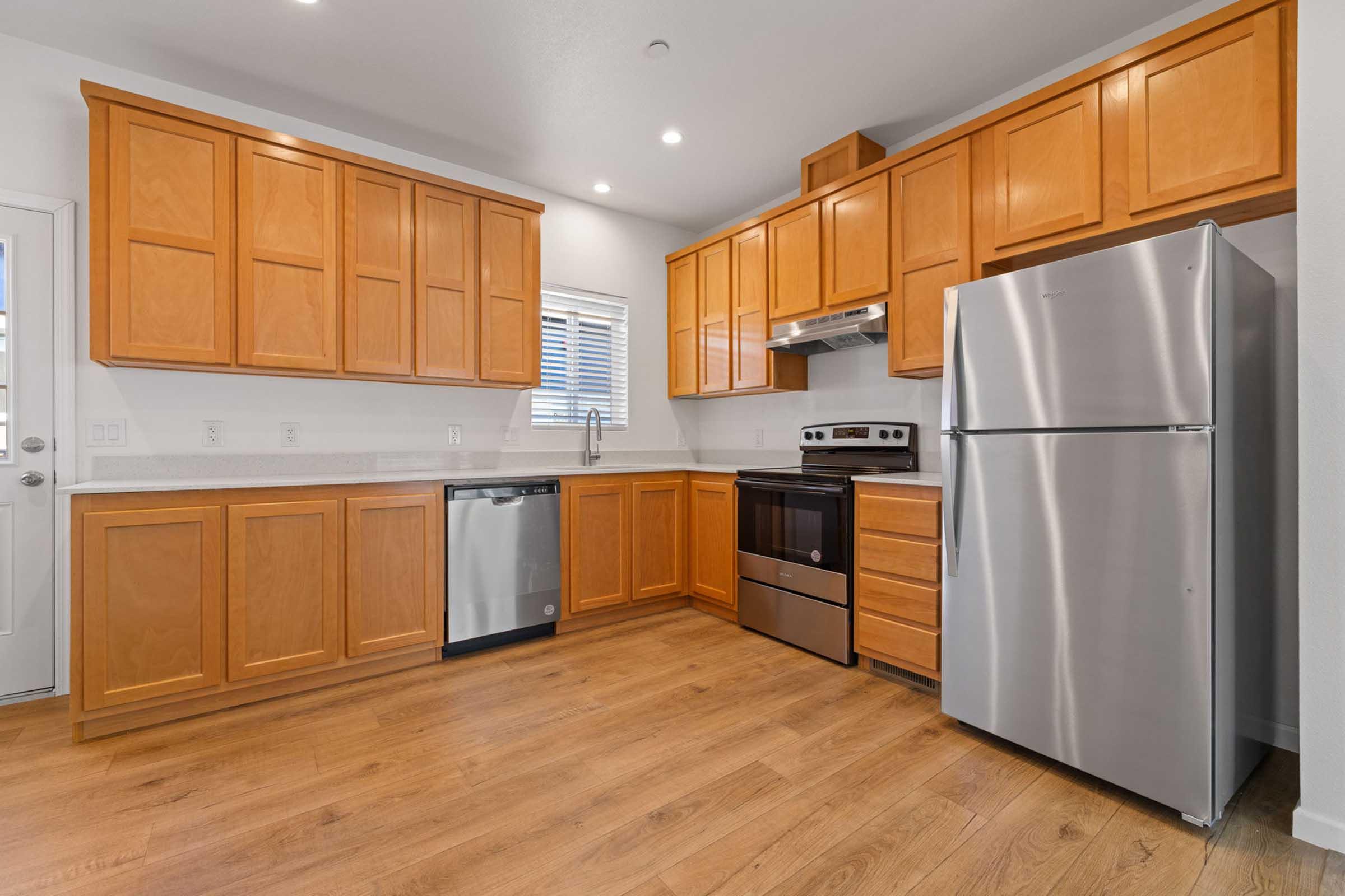 Modern kitchen featuring wooden cabinetry, stainless steel appliances including a refrigerator, dishwasher, and oven, along with a sleek countertop. Bright lighting enhances the warm tones of the wood and the spacious layout. A window allows natural light to enter.