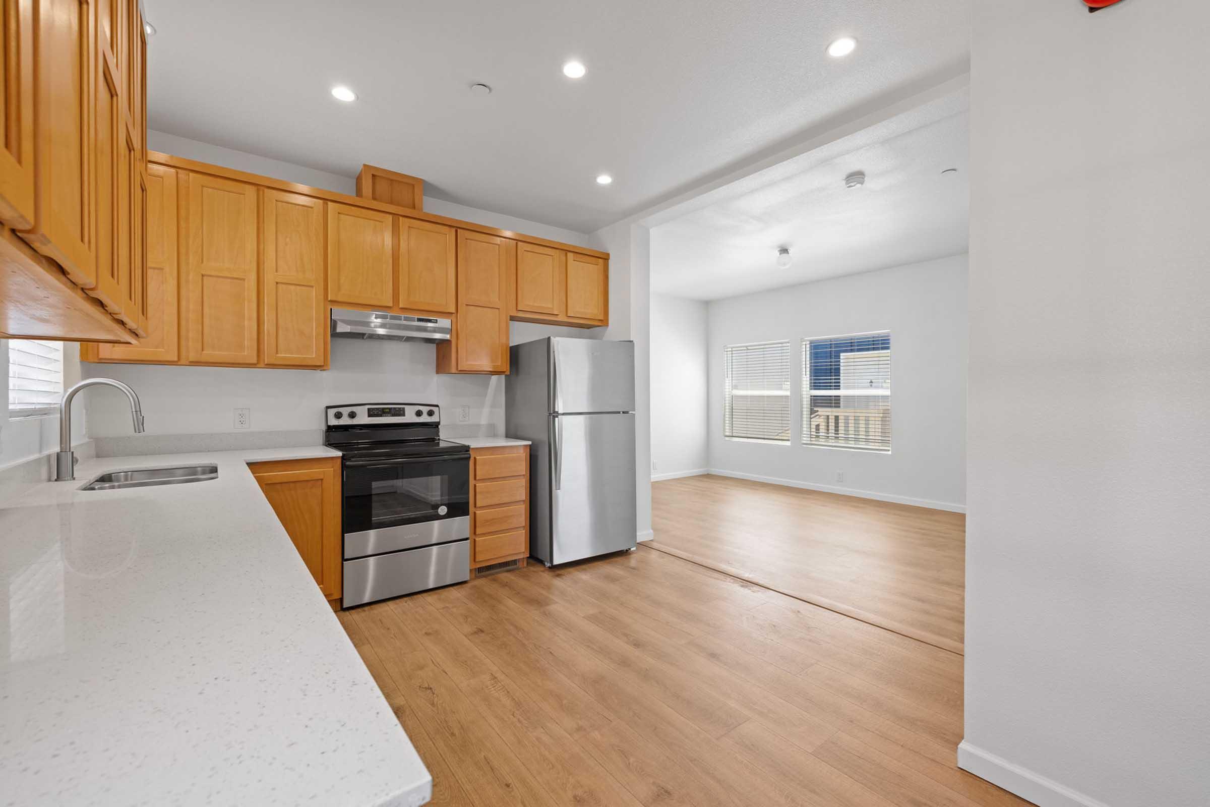 Modern kitchen with wooden cabinets, stainless steel appliances, and a white countertop. The layout features a spacious area connecting to a well-lit living space with large windows. The floor is finished with light wood, creating a warm and inviting atmosphere.