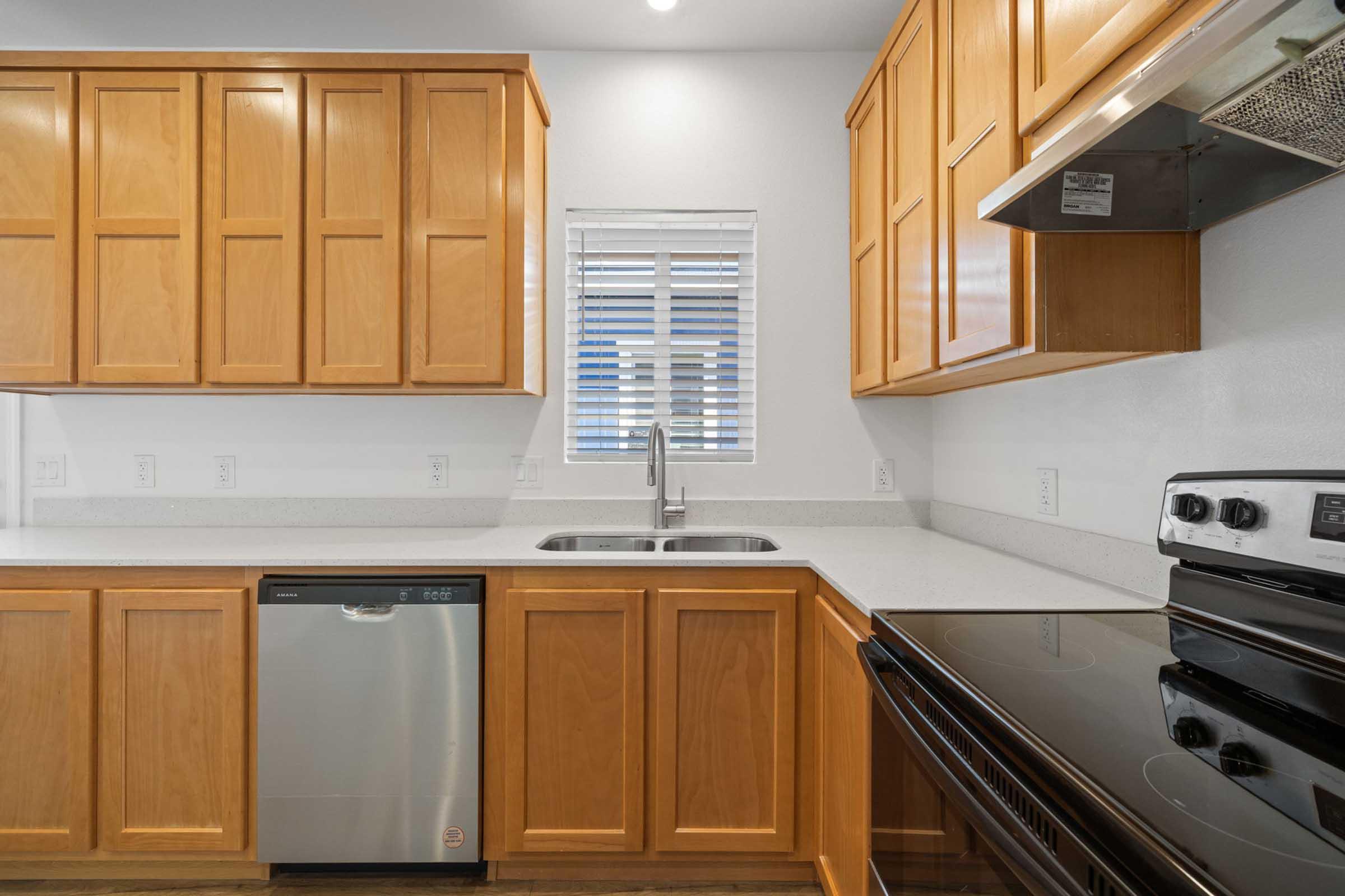 Modern kitchen featuring wooden cabinets, a stainless steel dishwasher, a sink with a faucet, and an electric stove. The countertop is light-colored, and there's a window with blinds above the sink, allowing natural light into the space.