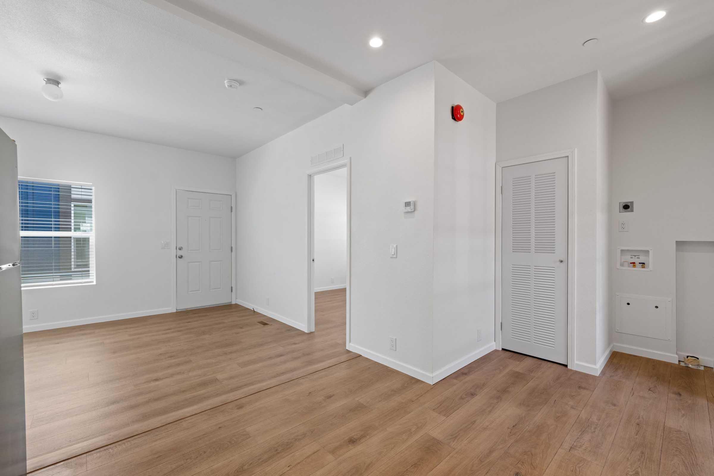 A bright, empty room with light-colored walls and wooden flooring. There is a door on the left, a white closet with louvered doors on the right, and a small wall-mounted panel. Recessed lighting and a smoke detector are visible on the ceiling, creating a modern, minimalist aesthetic.