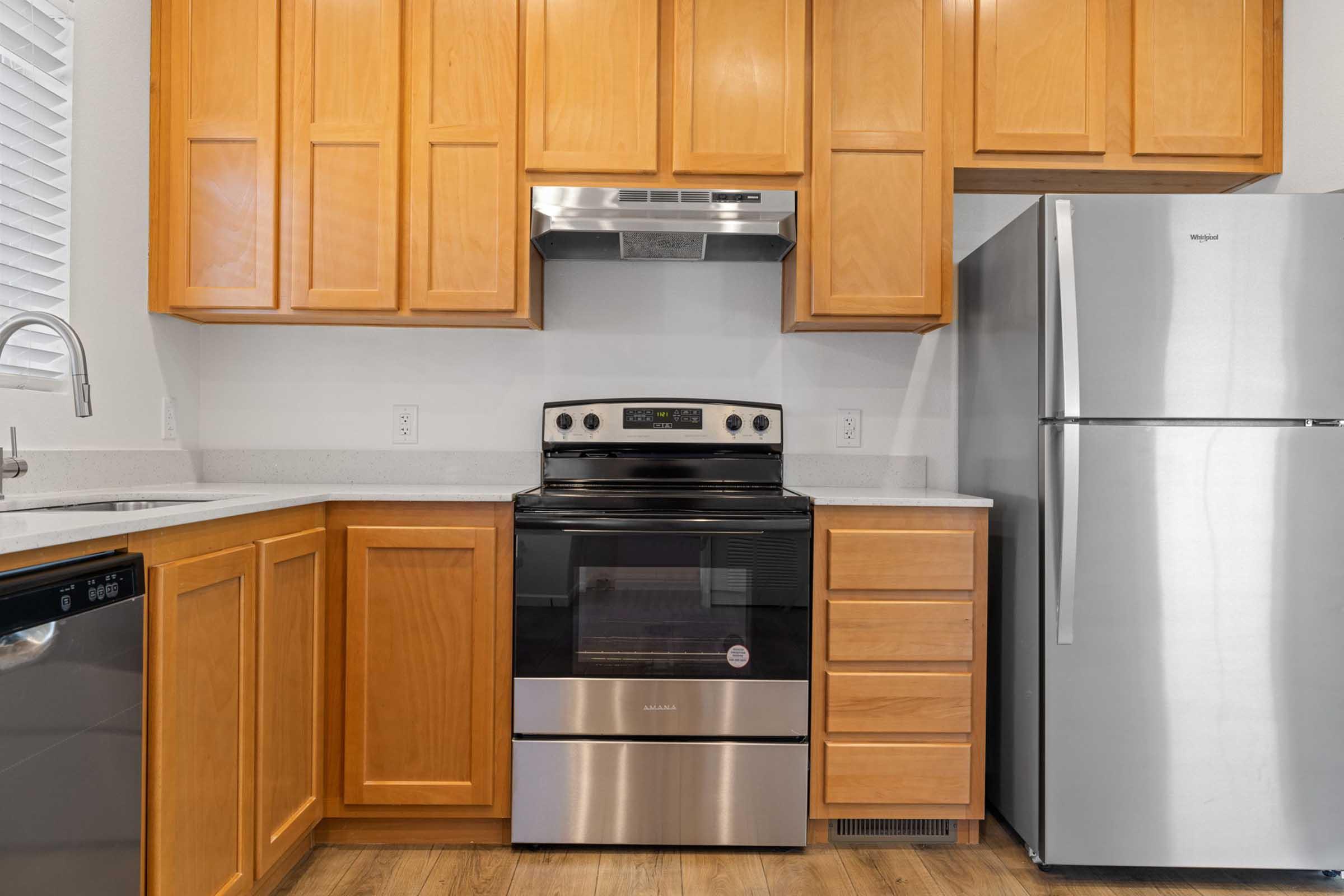 Modern kitchen with wooden cabinets, a stainless steel refrigerator, an electric oven with a black and stainless steel finish, and a silver dishwasher. Countertops are light-colored with a subtle texture. The space is well-lit with natural light coming through a window.