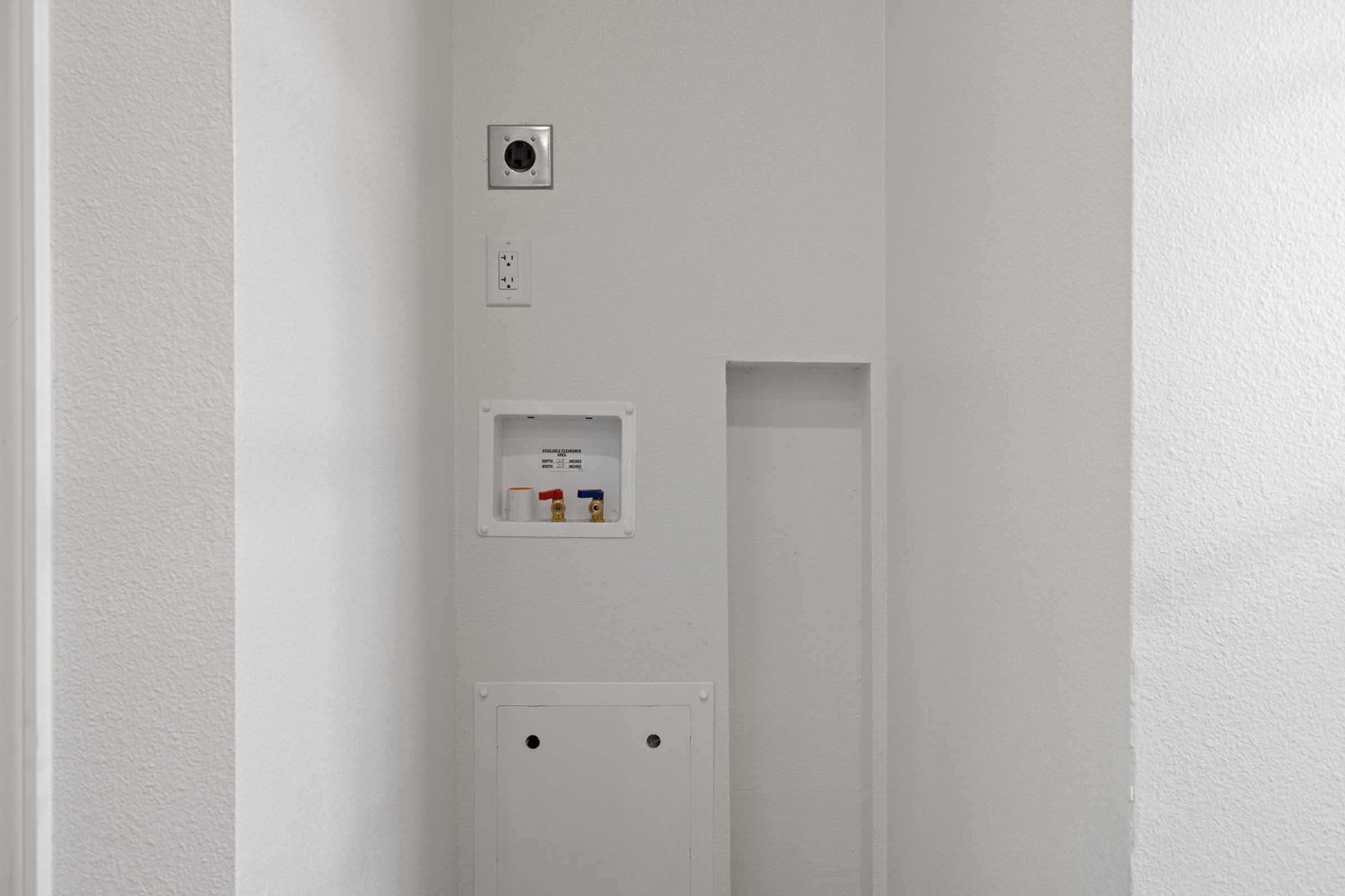 A clean, minimalist interior wall painted white, featuring a small electrical panel with circuit breakers, a smoke detector on the ceiling, and a recessed area. The design emphasizes simplicity and functionality in a well-lit, uncluttered space.