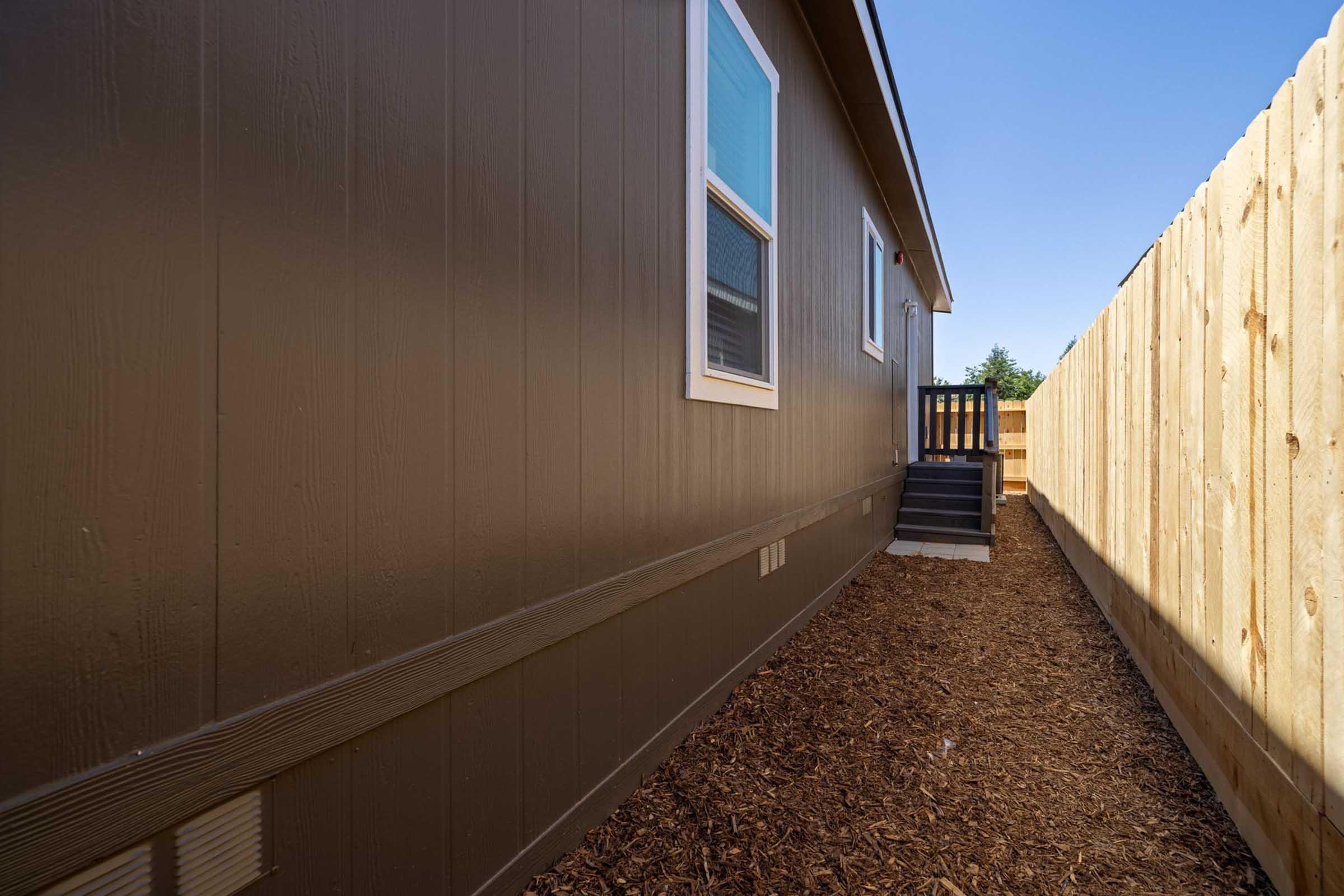 Narrow pathway beside a brown-painted house leading to a set of steps. The area is lined with a wooden fence and features mulch along the ground. The sky is clear and blue, creating a bright atmosphere.